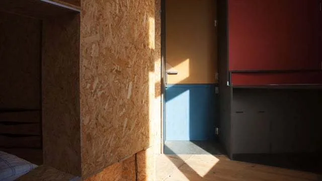 An interior space features textured OSB wall panels, a yellow and blue color-blocked door, and sharp shadows cast across the light wood floor.