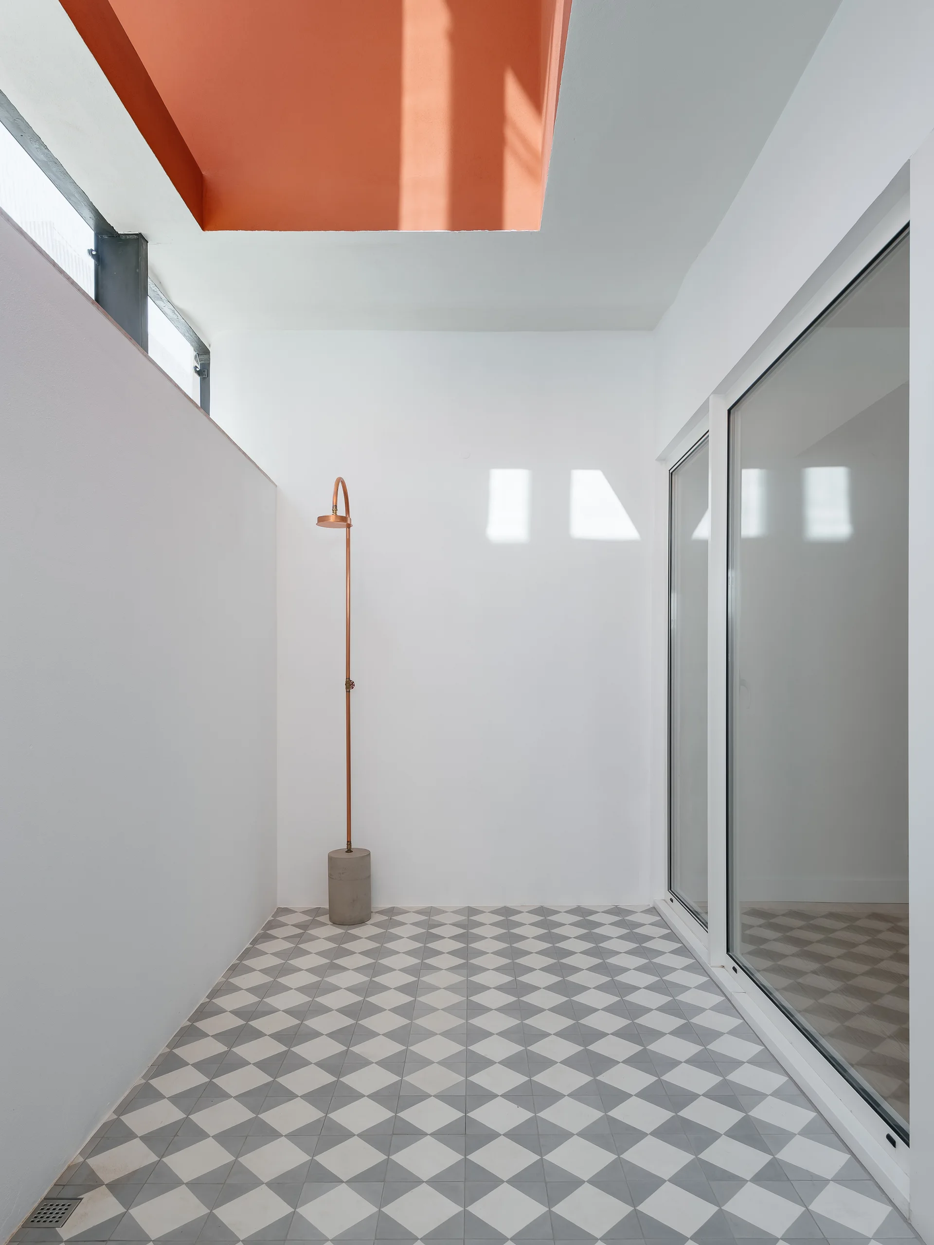 A minimalist outdoor shower space with a freestanding copper fixture, grey and white geometric tile flooring, and a terracotta-colored ceiling recess.