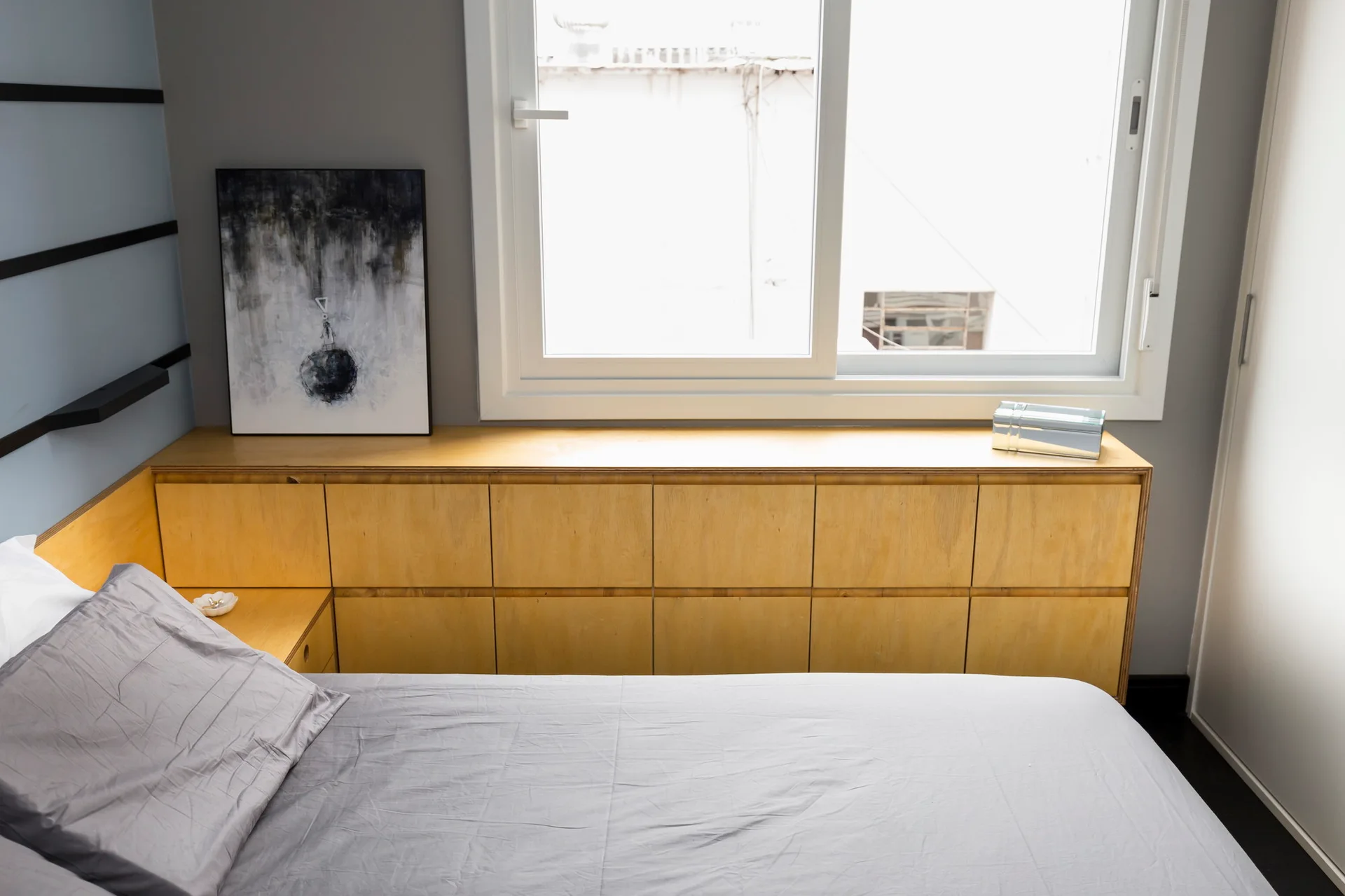 A minimalist bedroom featuring a light wood built-in storage cabinet under a window next to a bed with grey sheets.