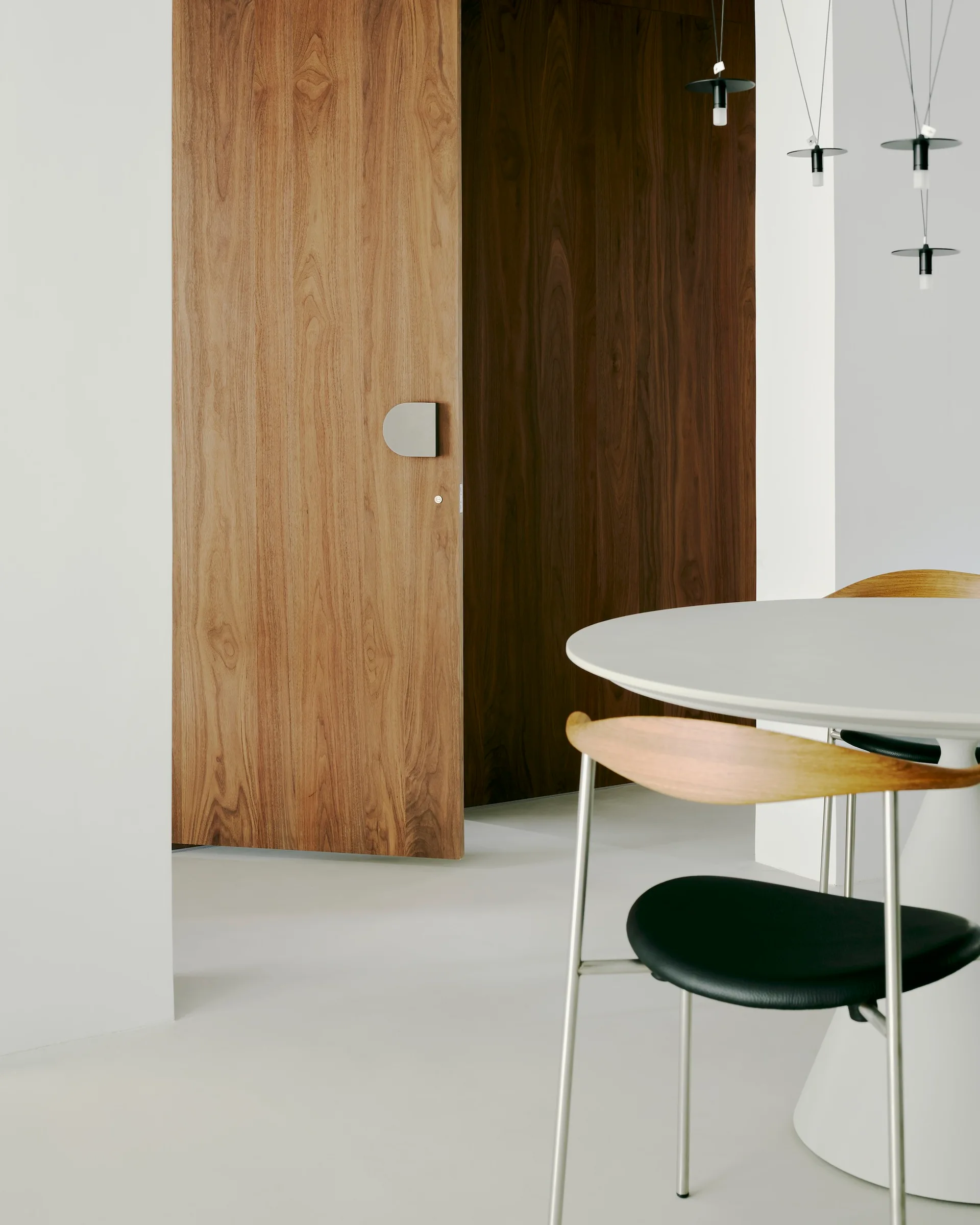 A white minimalist dining room features a round white table, a wood-backed chair, and a warm wood door with a unique metallic handle.