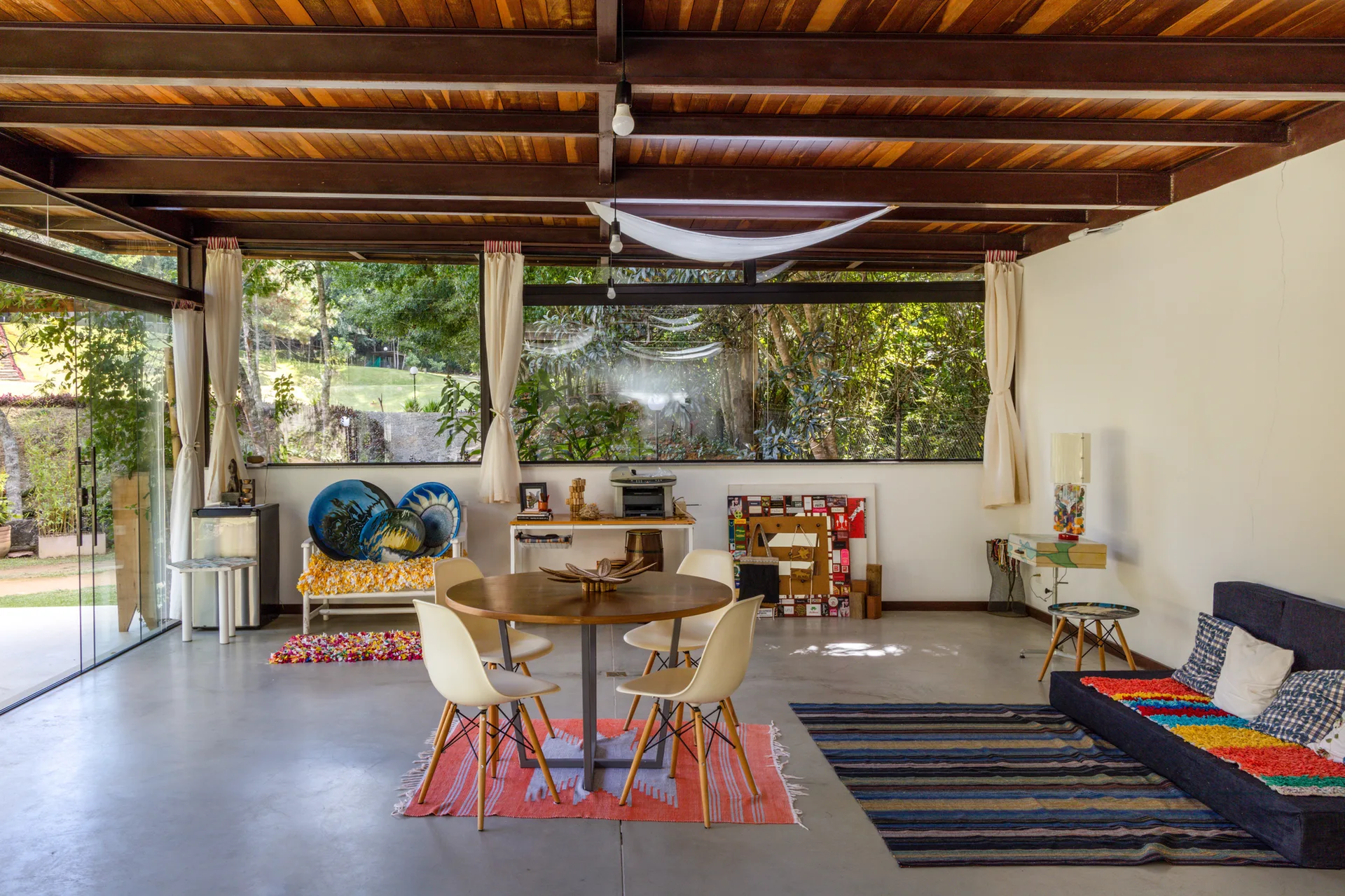 A spacious open-concept room features a round wooden dining table, white chairs, a low sofa, and an exposed timber ceiling next to glass walls.