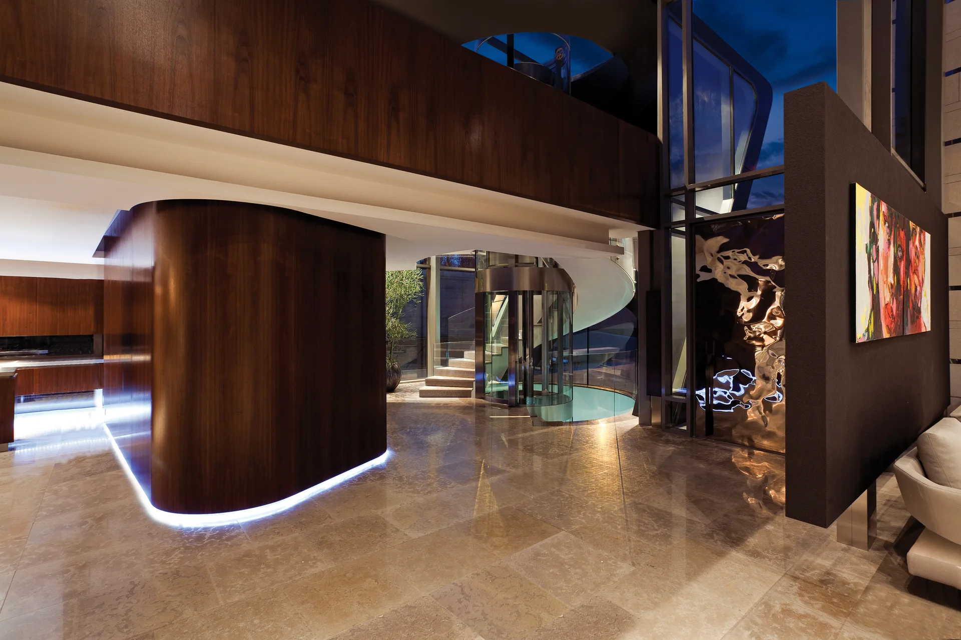 A modern entryway featuring a curved dark wood partition wall, a cylindrical glass elevator, and highly polished stone floor tiles.