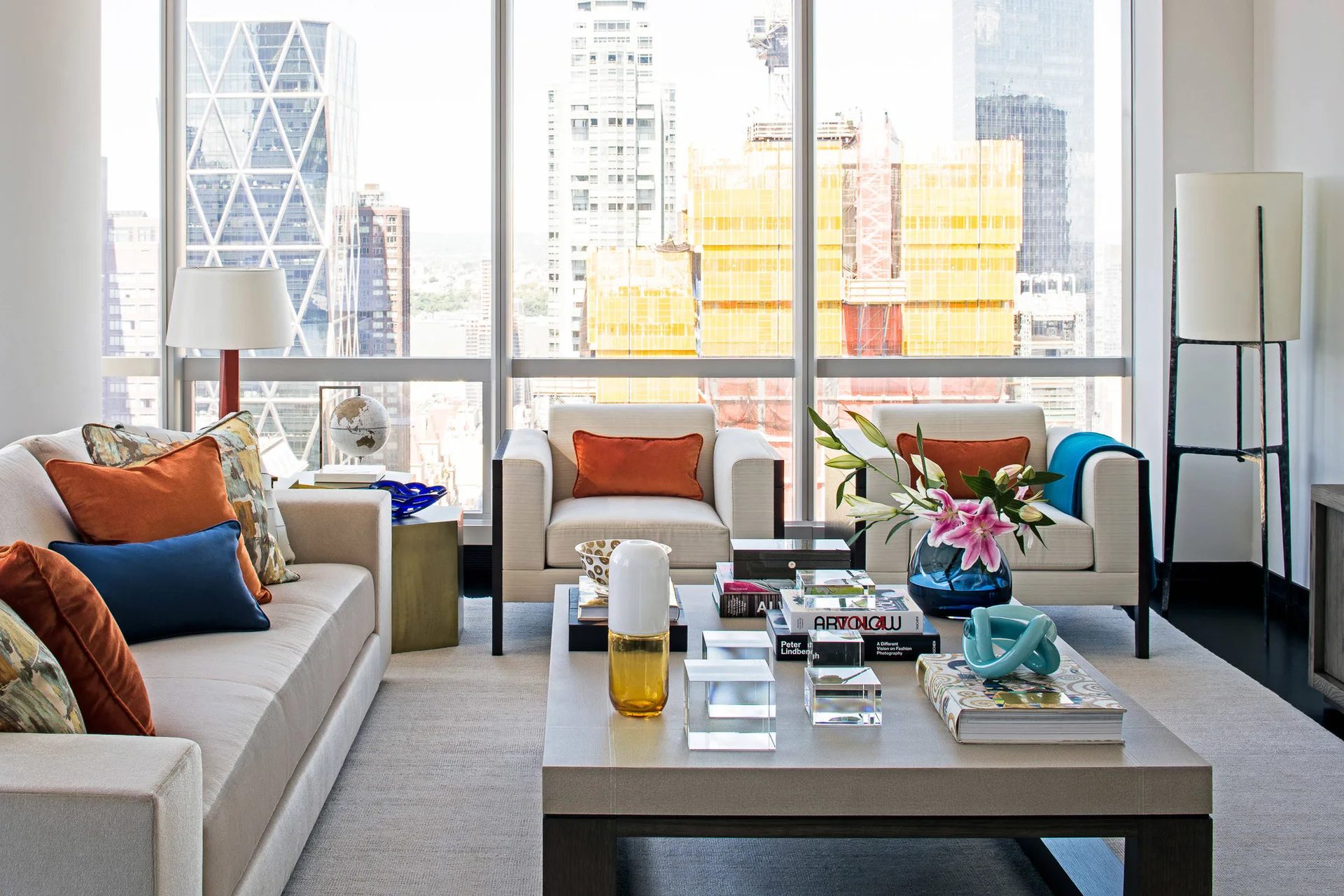 A modern high-rise living room features a beige sofa and armchairs decorated with orange and blue pillows around a low coffee table with a city view.