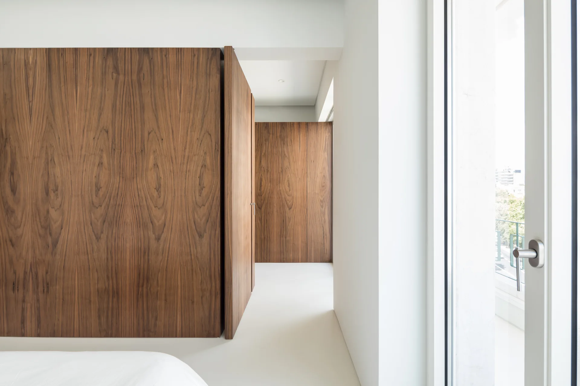 A minimalist white hallway featuring floor-to-ceiling wood-grained panels with a partially open integrated door next to a glass patio door.