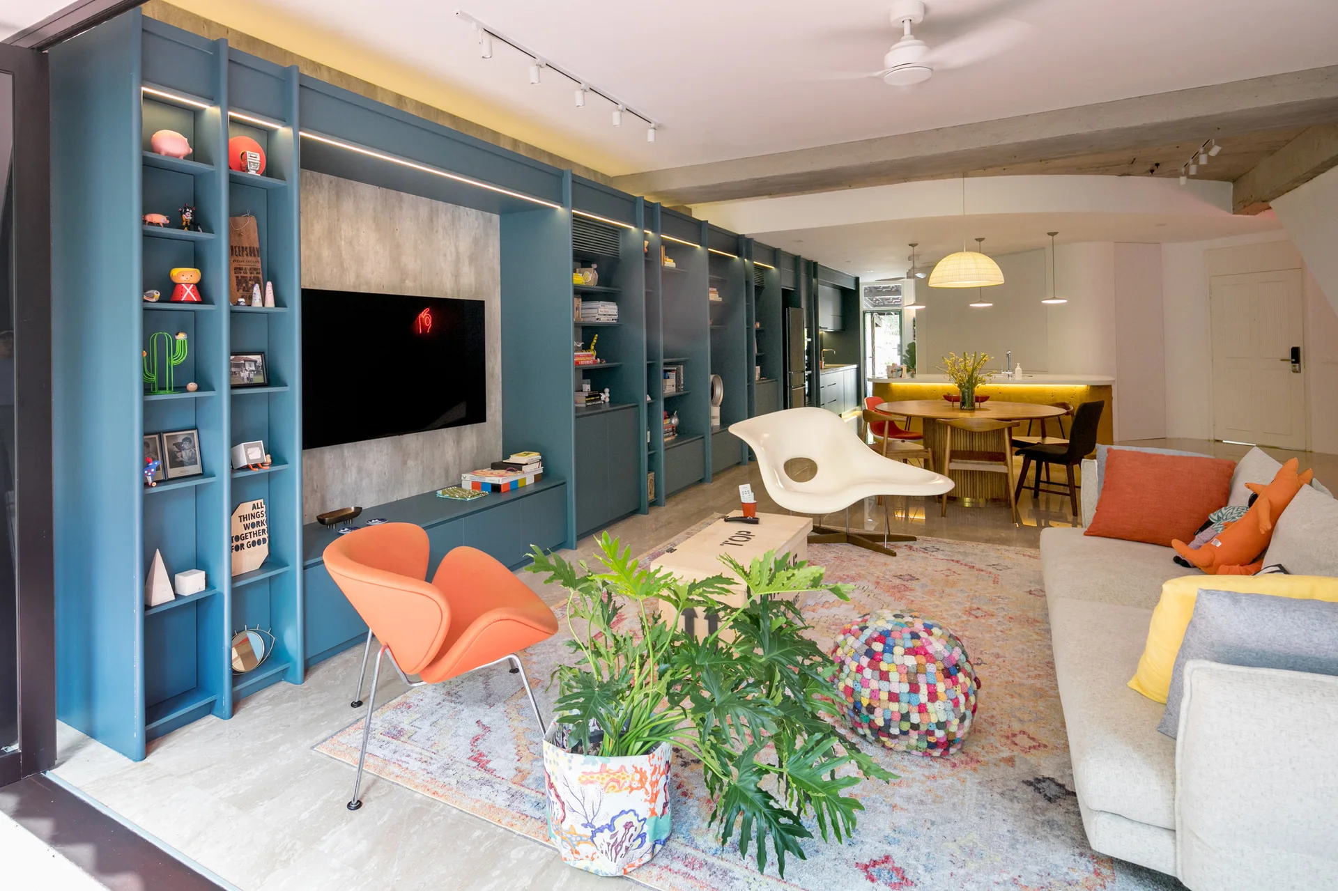 A modern living room features a large blue built-in shelving unit, a cream-colored sofa with orange accent pillows, and an orange modern armchair.