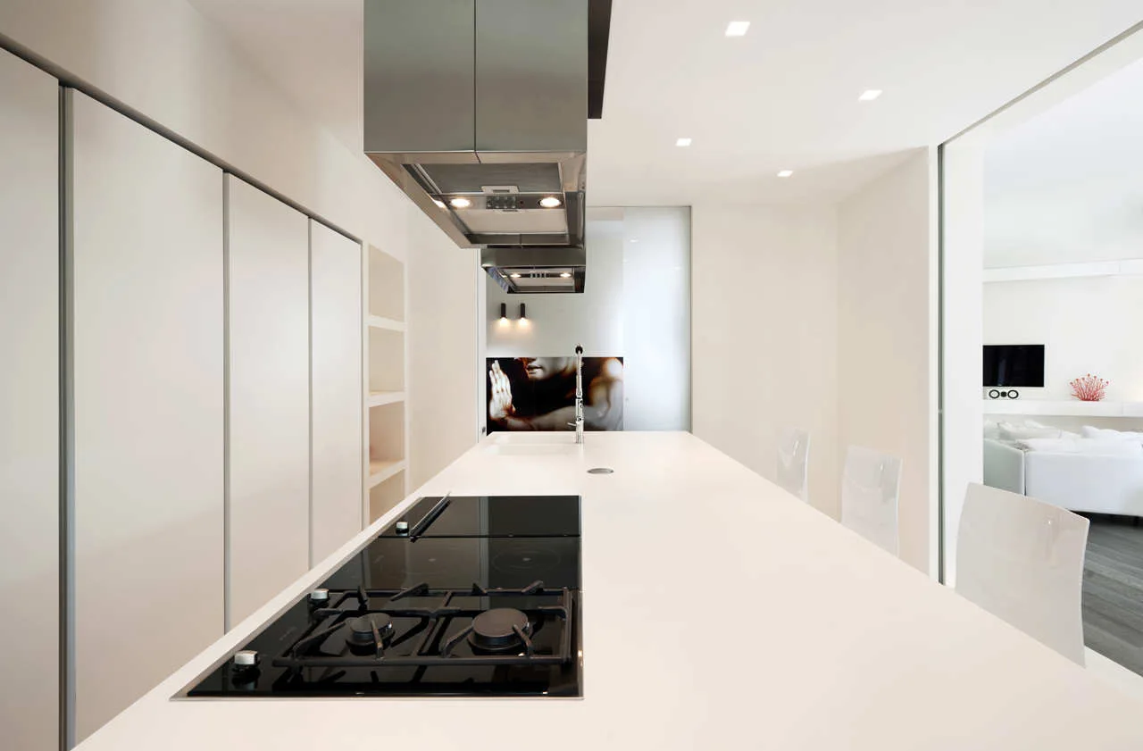 A long minimalist white kitchen island with a black cooktop and stainless steel range hood next to floor-to-ceiling white cabinets.