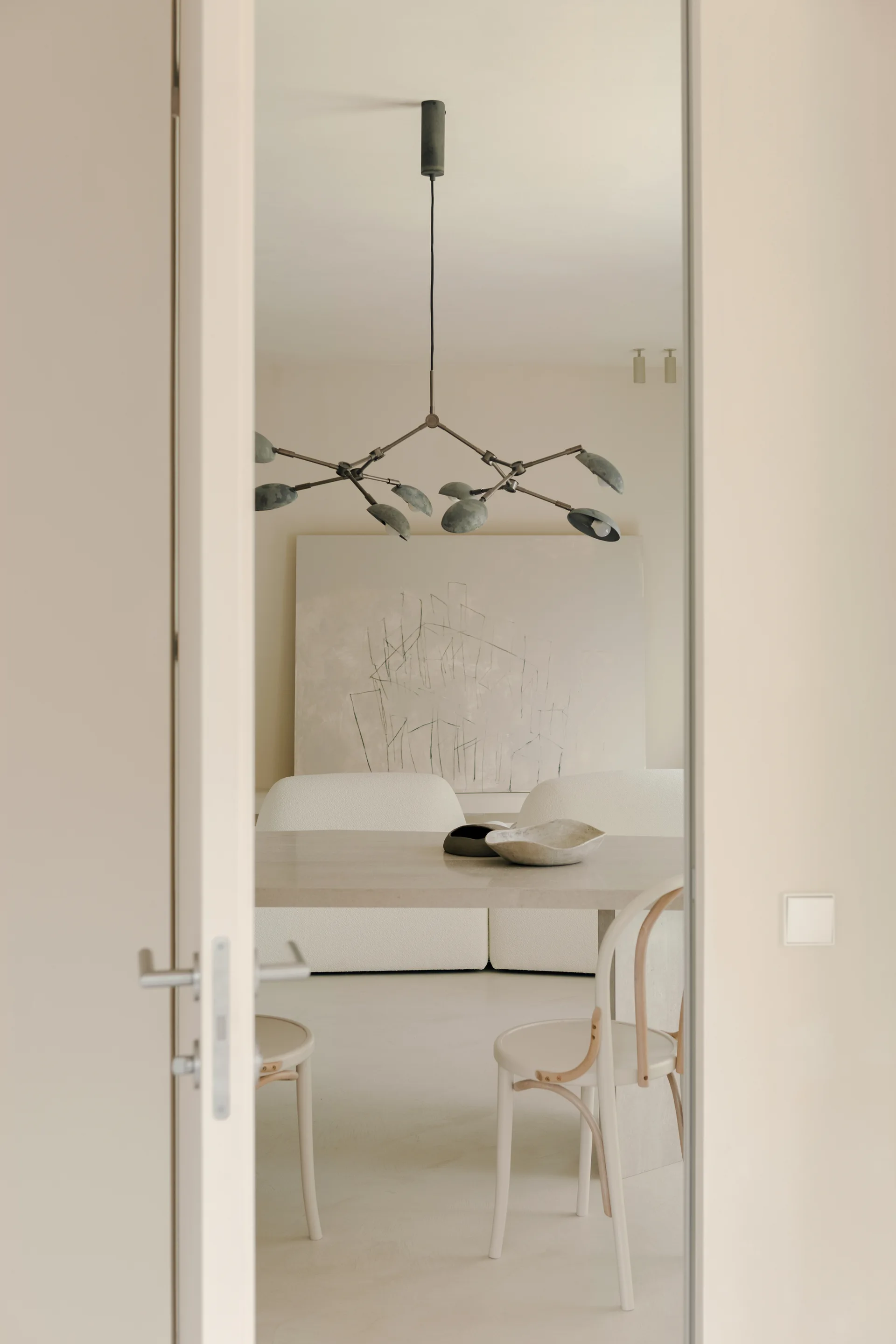 A view through a doorway into an off-white dining area with a wood table, curved chairs, a branching metal chandelier, and a curved cream sofa.