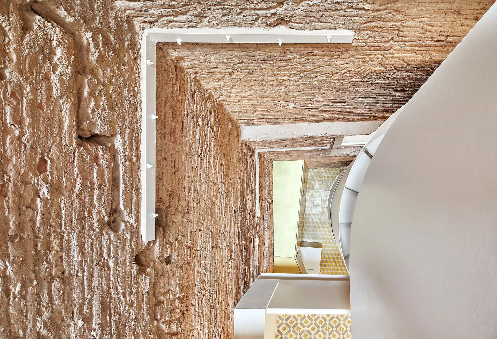 A high-angle architectural perspective looking down a narrow stairwell with rough brick walls and a curved white staircase structure.