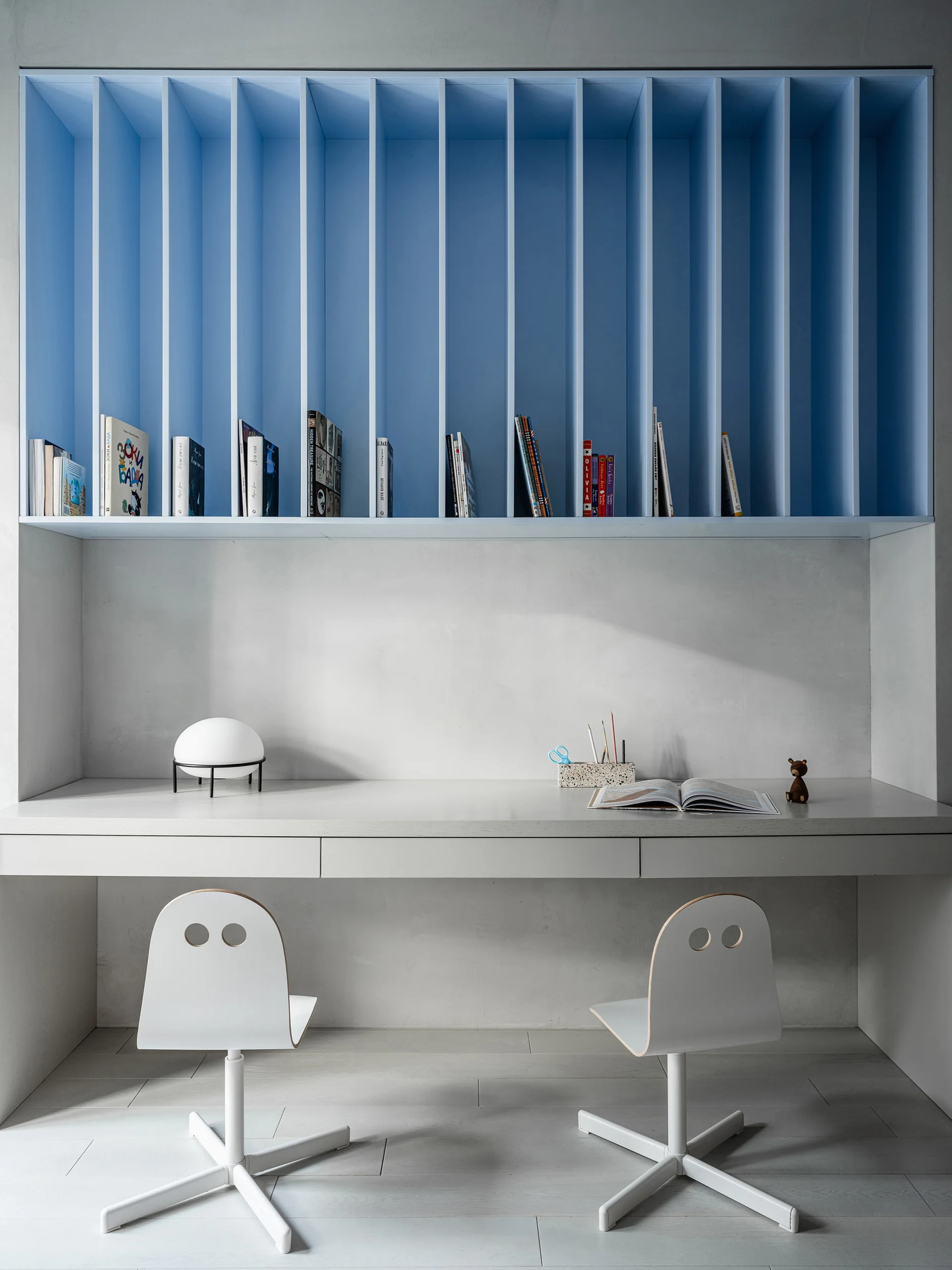 A minimalist grey study alcove features a built-in desk with two white chairs beneath a striking blue shelving unit with vertical dividers.