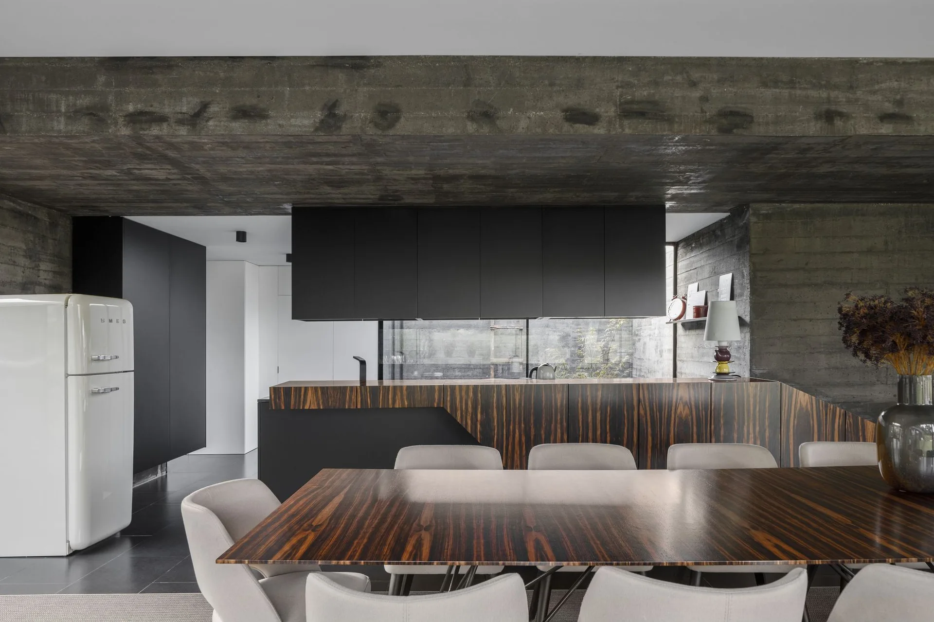 A modern dining area features a striped wood table and a kitchen island with matching wood veneer under an exposed concrete ceiling.