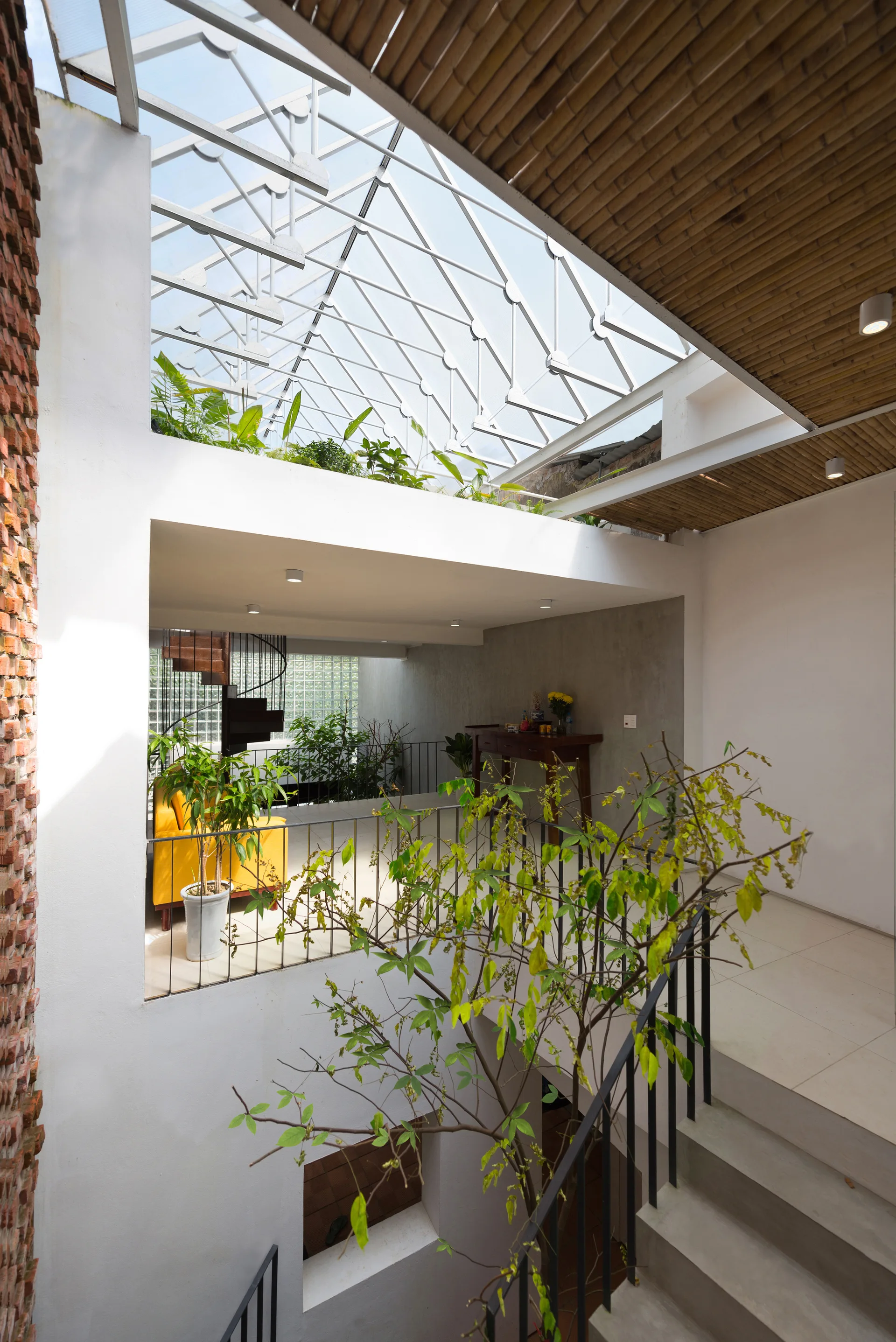 A multi-level modern interior featuring a concrete staircase, a central atrium with a glass skylight roof, indoor plants, and an exposed brick wall.