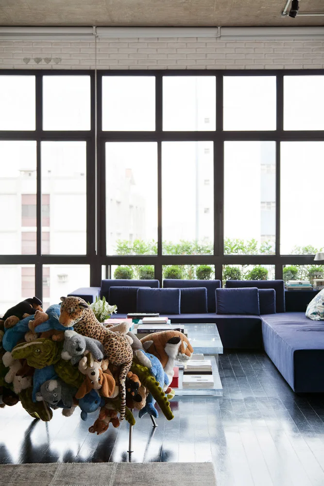 A modern loft living room with a navy blue sectional sofa and an accent chair made of plush toys in front of large windows.