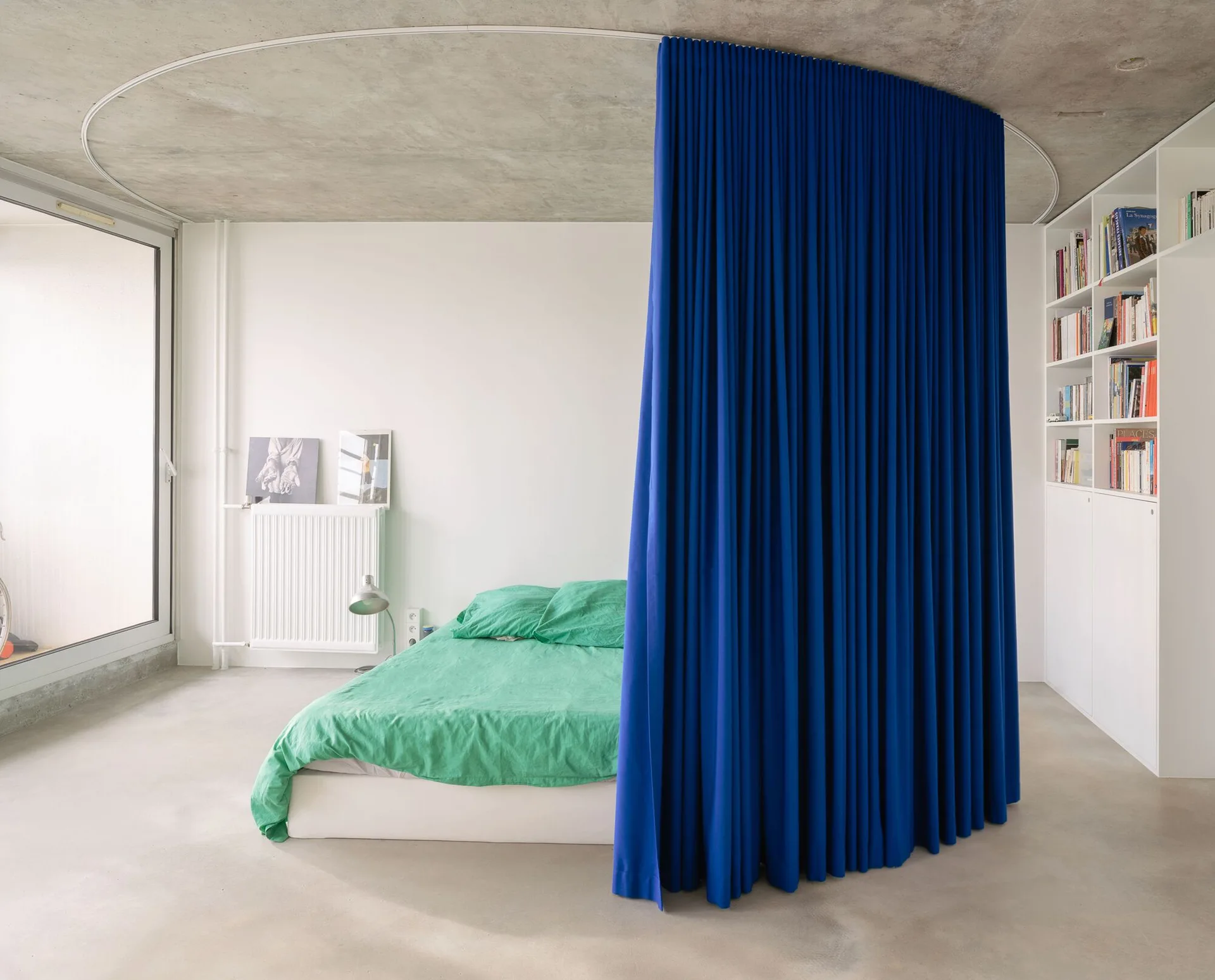 A modern bedroom features a low bed with green linens, a curved blue partition curtain on a ceiling track, and a raw concrete ceiling.
