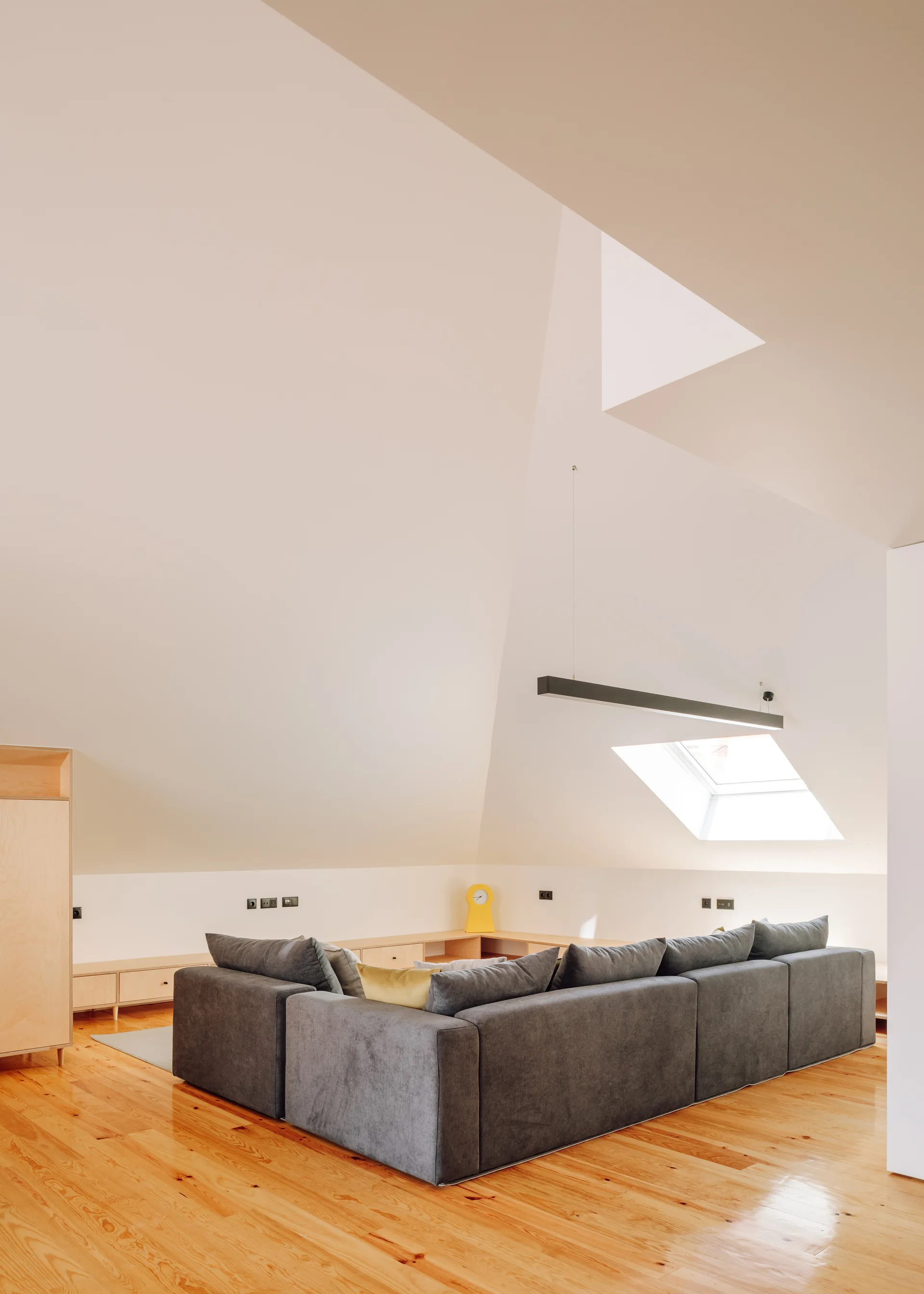 An attic living room with a large grey sectional sofa, low light wood cabinets, and a sloped white ceiling with a skylight.