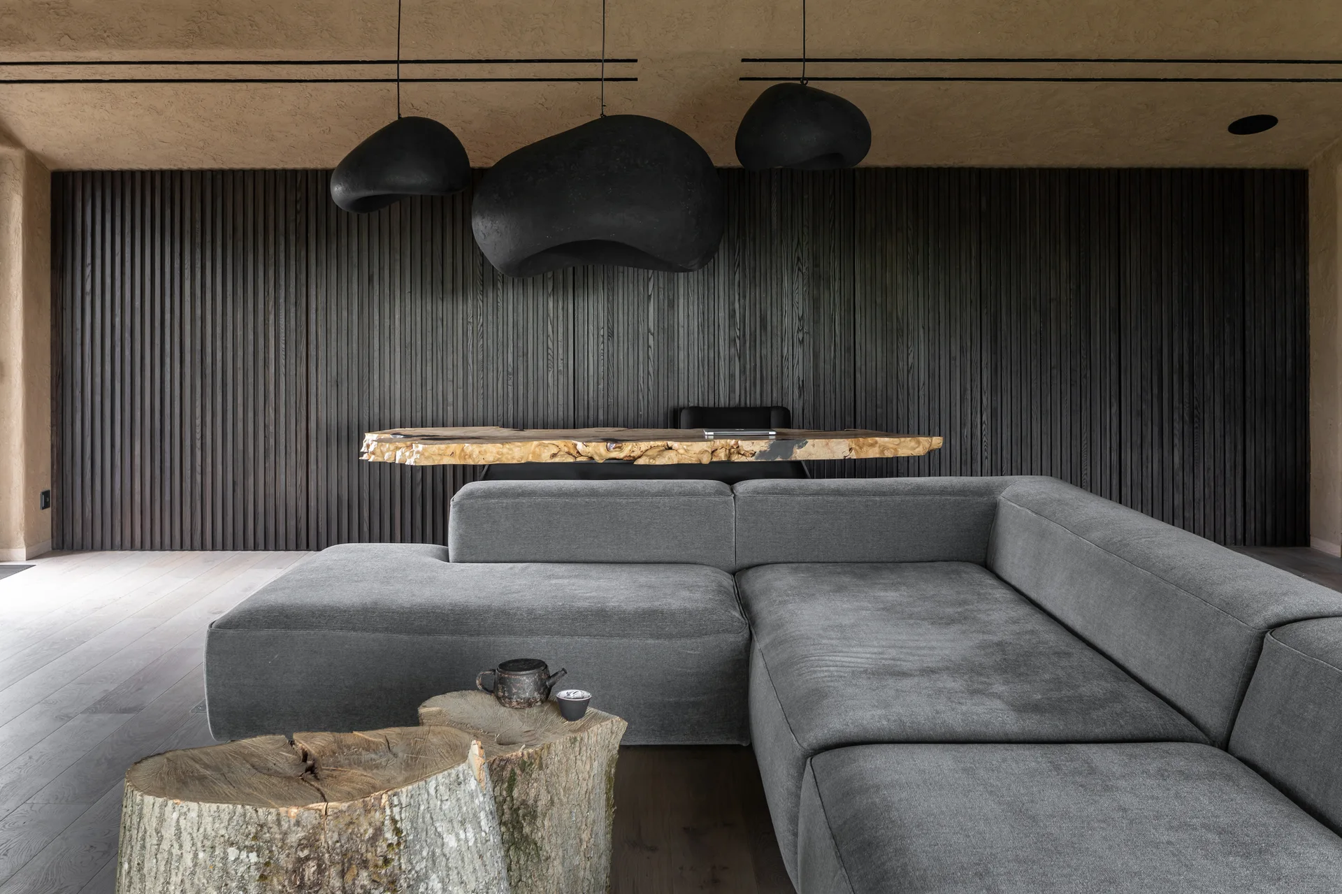 A grey sectional sofa sits in a living room with raw wood log coffee tables, a dark slatted accent wall, and three black sculptural pendant lights.