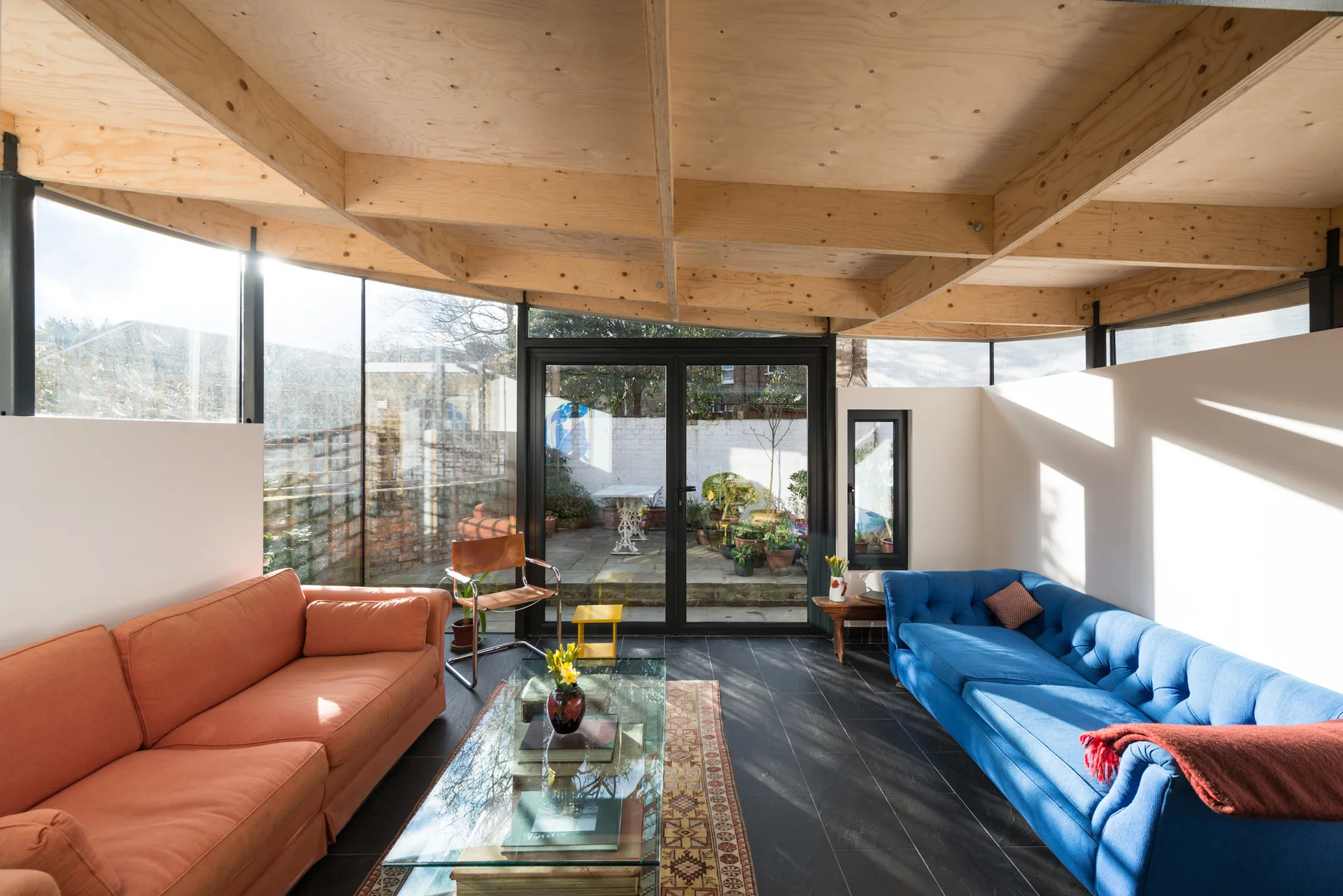 A sunlit living room featuring an orange sofa and a blue sofa facing each other under an exposed plywood ceiling, with glass walls looking out to a garden.