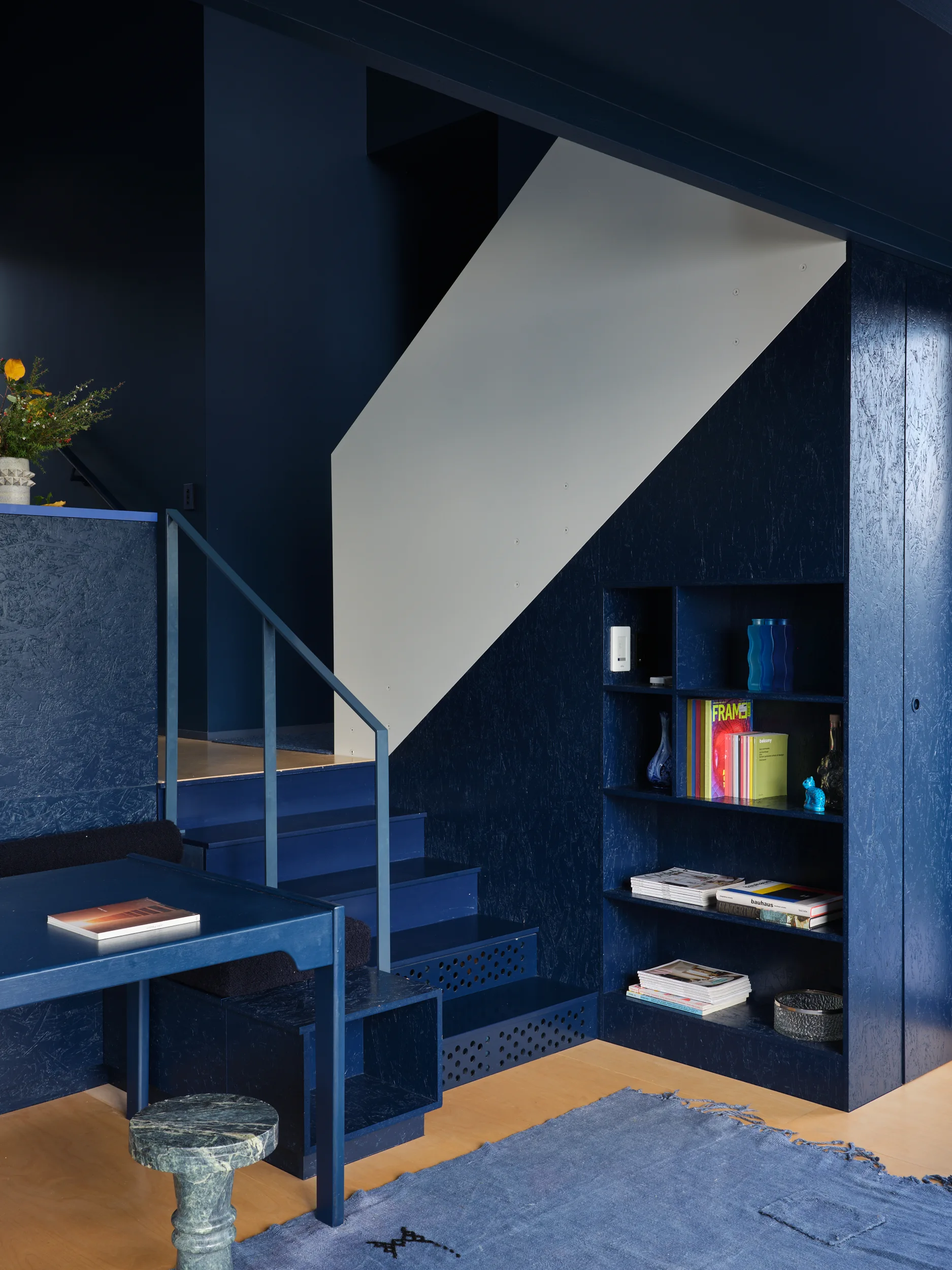 A deep blue staircase made of textured painted wood features integrated open shelving, perforated lower steps, and a matching blue table.