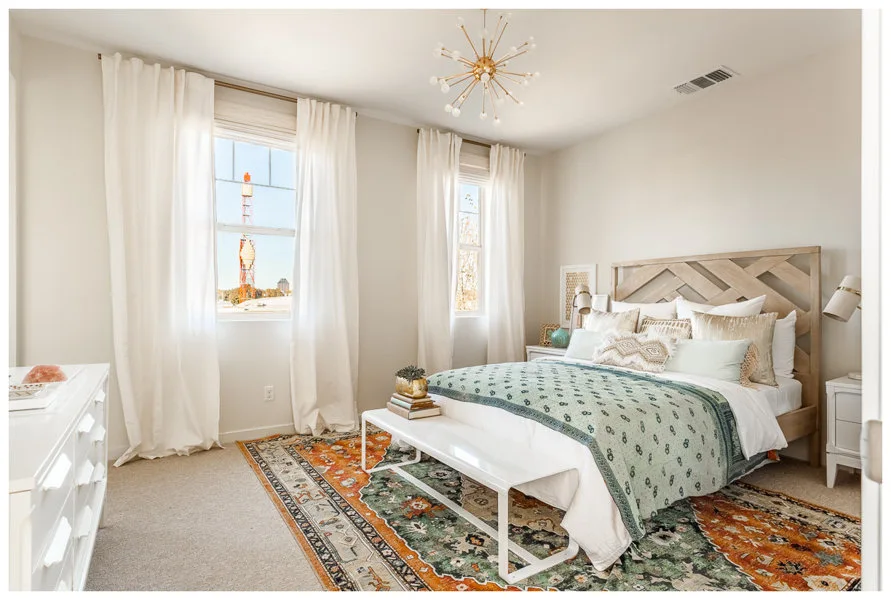 A bedroom features a light wood herringbone headboard, a white bench at the foot of the bed, and a colorful patterned area rug.