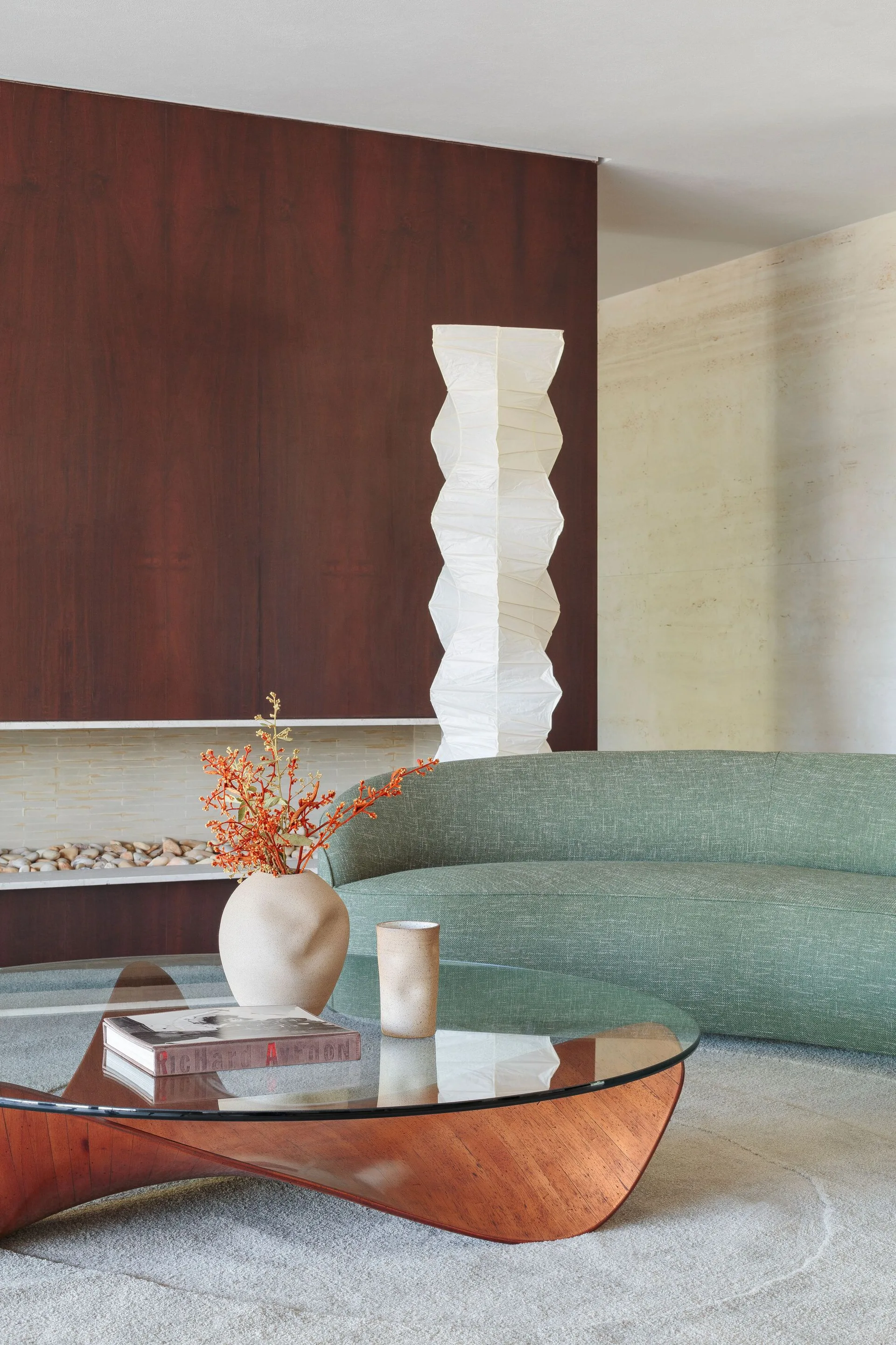 A modern living room features a curved green sofa, a glass-top coffee table with a sculptural wood base, a tall paper floor lamp, and a dark wood accent wall.