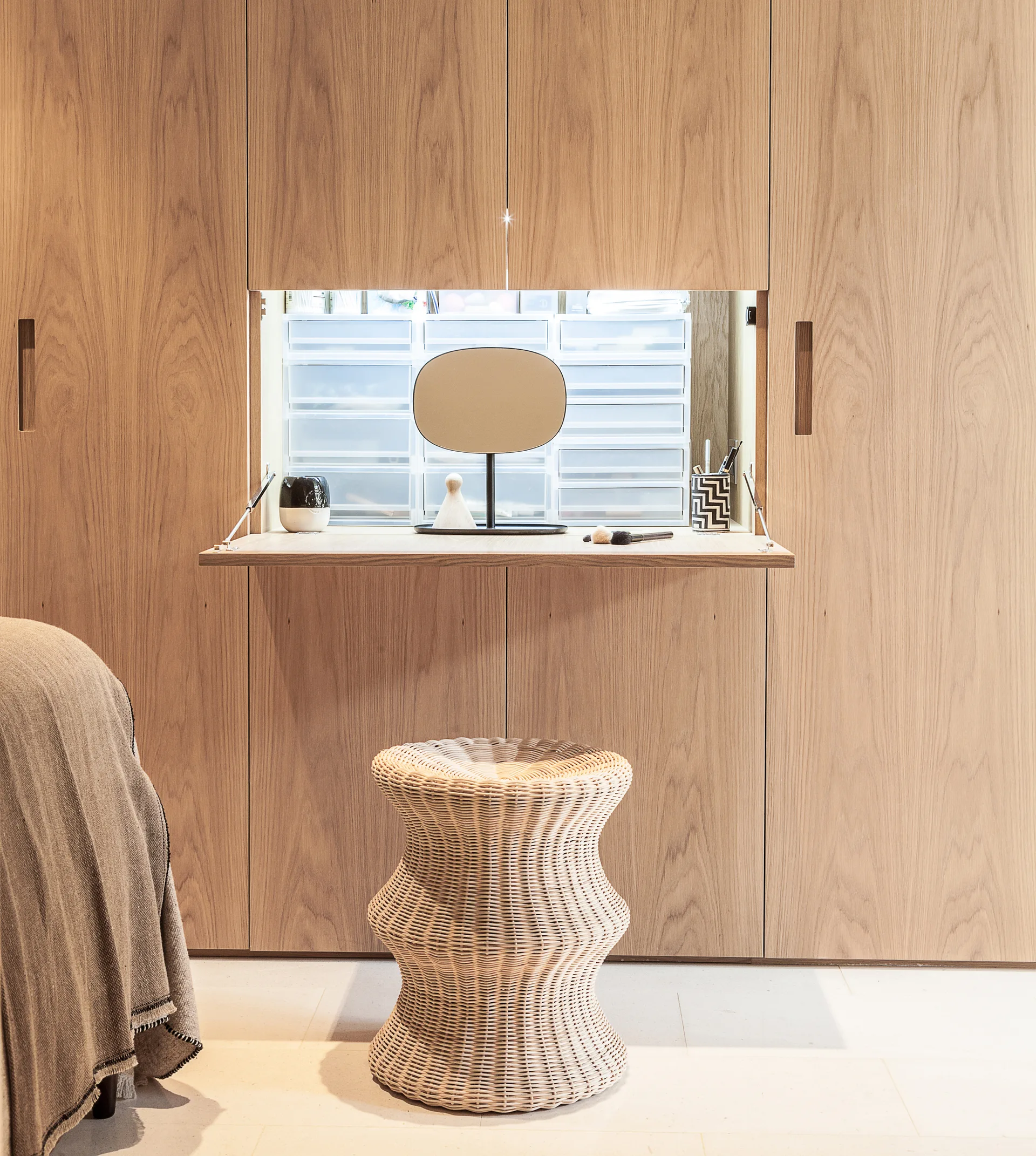 A light wood closet wall features a fold-down vanity table holding a small mirror and cosmetics, paired with a woven rattan hourglass stool.