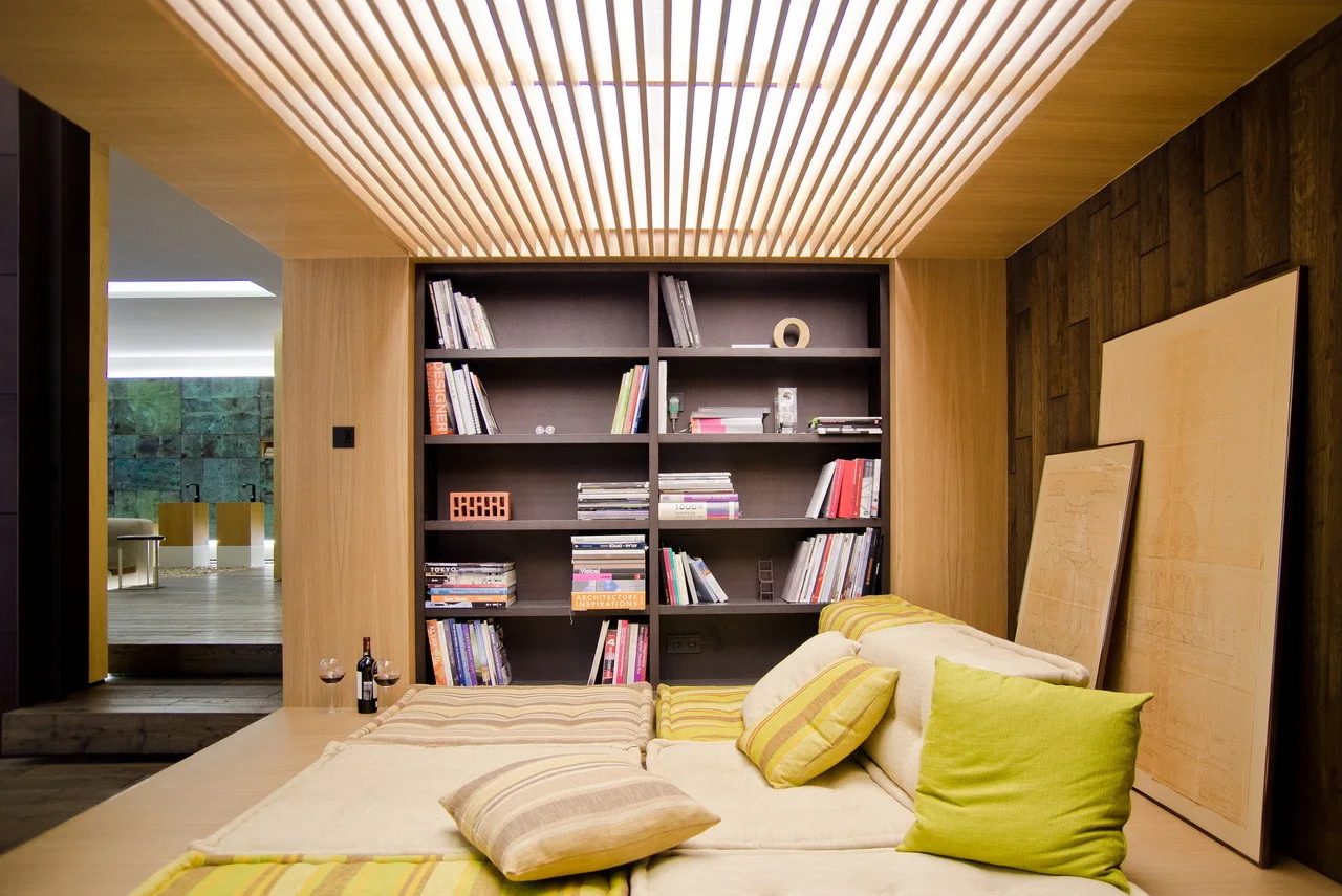 A low-profile platform bed sits beneath a lit wood slatted ceiling and in front of a dark built-in bookshelf.