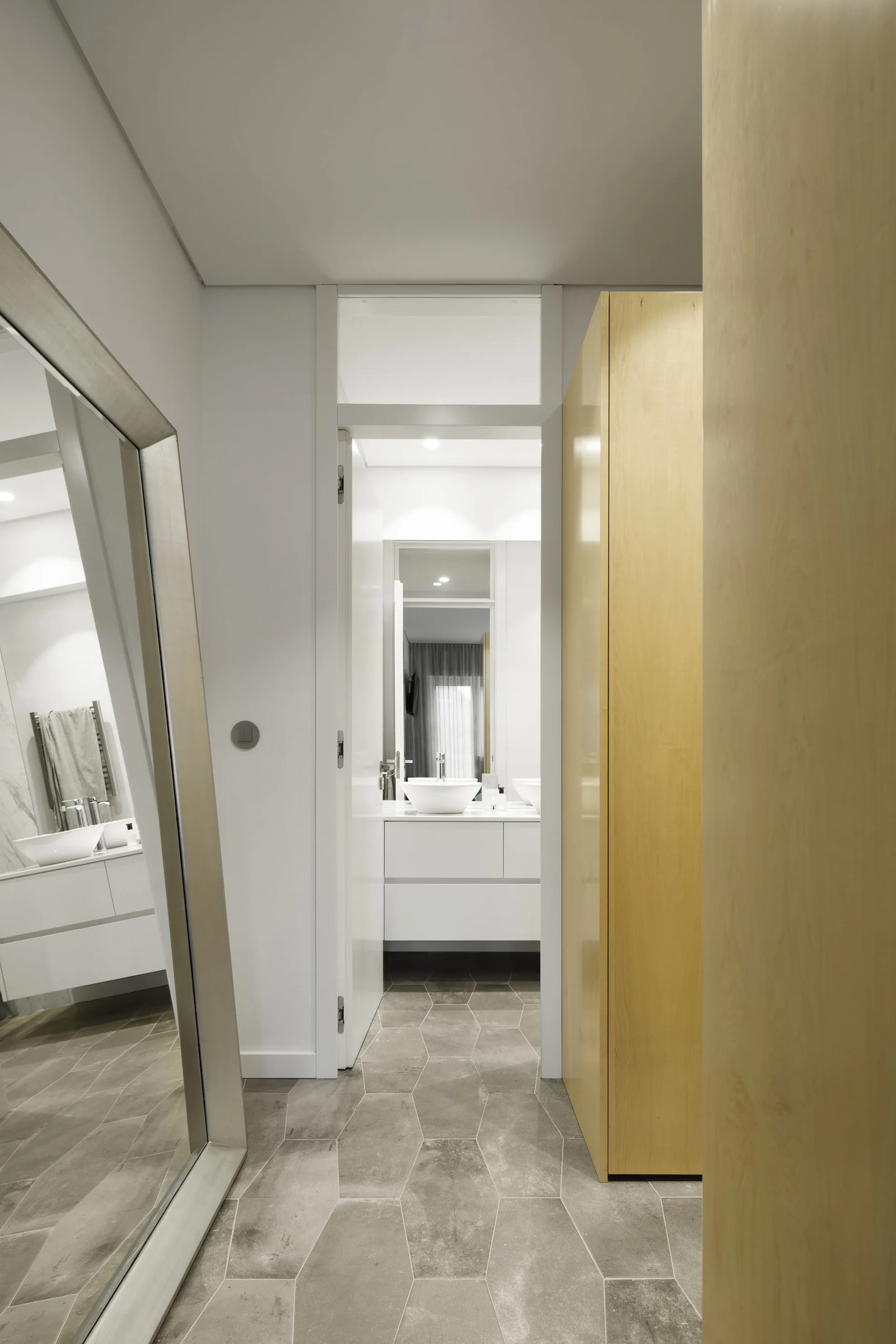 An entryway view featuring gray hexagonal floor tiles, a tall light wood cabinet, and a large mirror reflecting a white floating vanity.