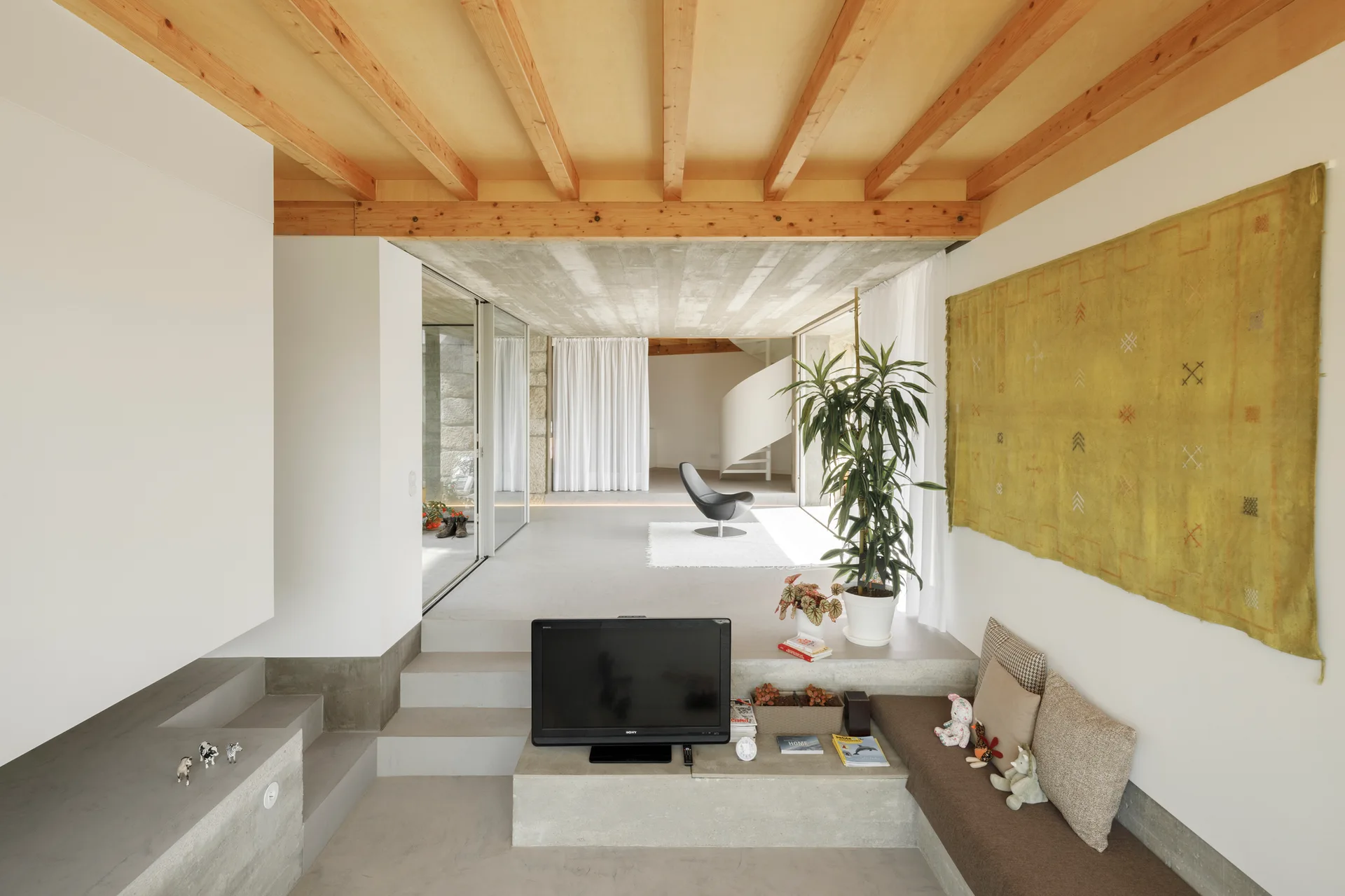 A sunken modern living room with a concrete floor, built-in bench seating, exposed timber ceiling beams, and a large yellow wall tapestry.