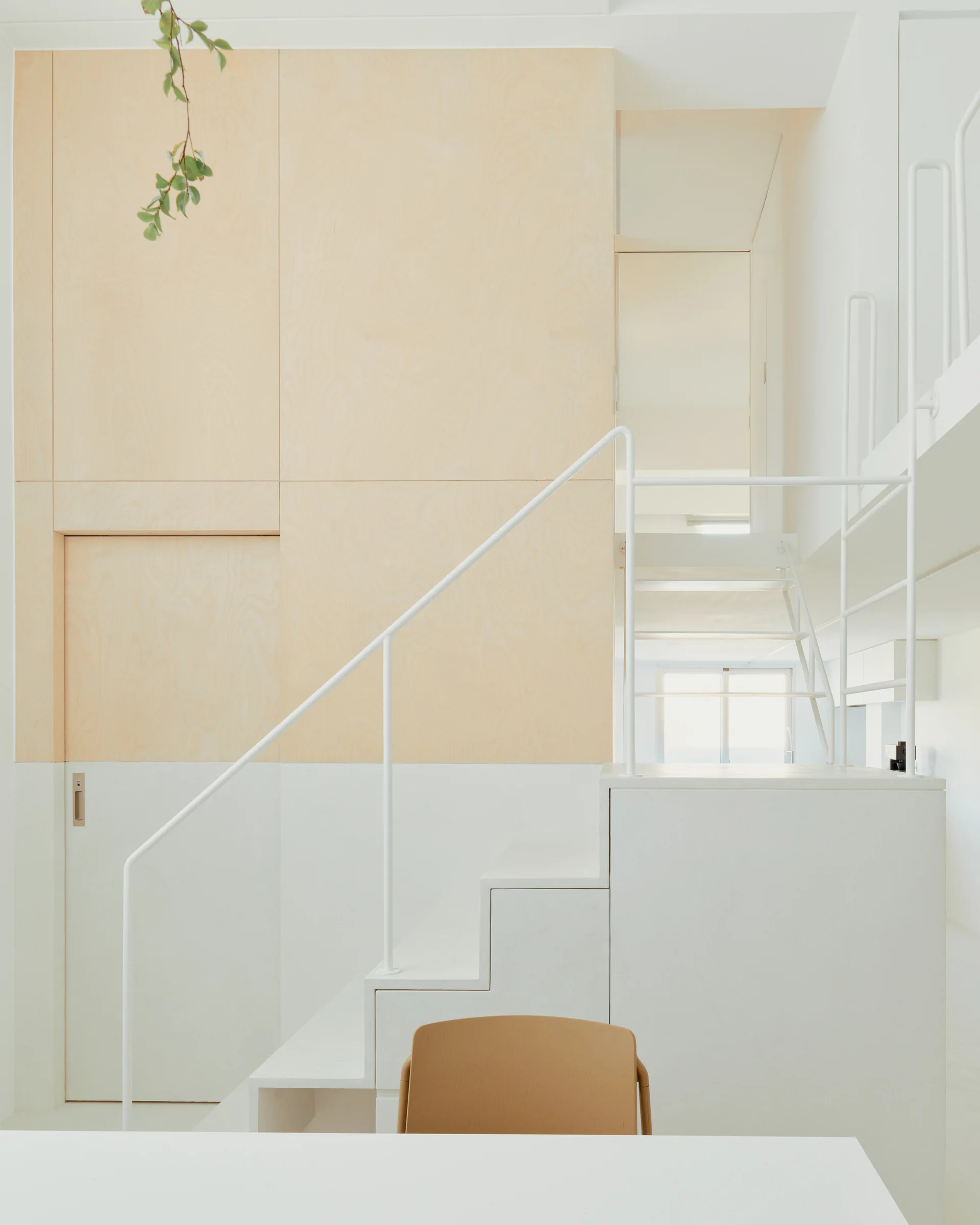 A minimalist white staircase with a thin metal handrail stands next to a two-tone wall of white paint and light plywood panels.