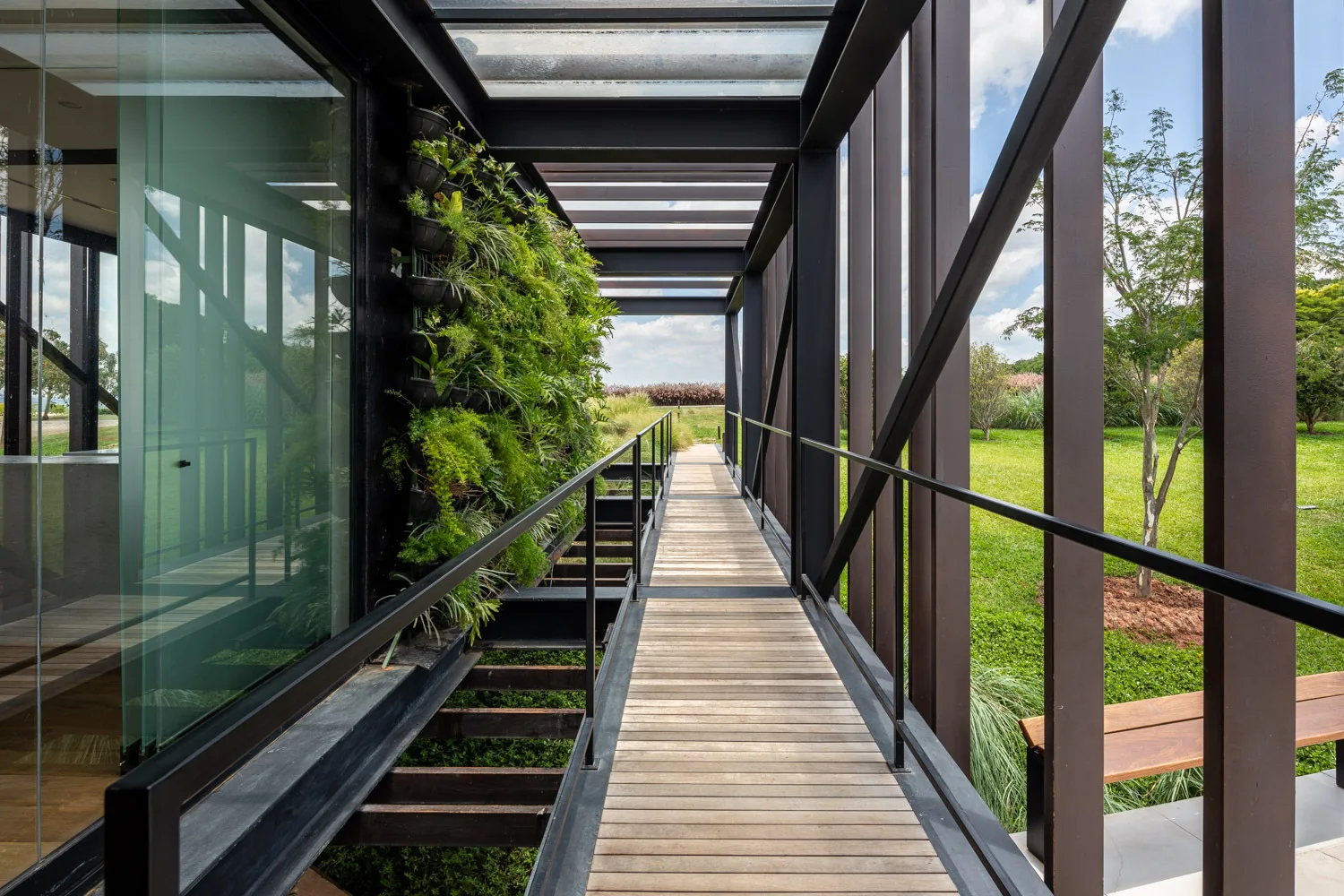A wooden plank walkway with black steel framing features a lush vertical garden wall on the left and vertical metal columns overlooking a green lawn.