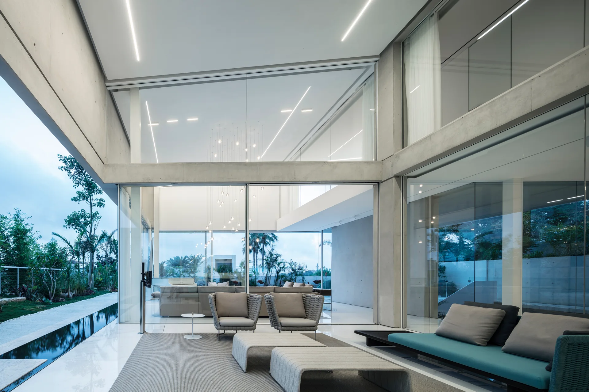 A minimalist living room features exposed concrete structural beams, polished white tile floors, woven grey armchairs, and floor-to-ceiling glass walls.