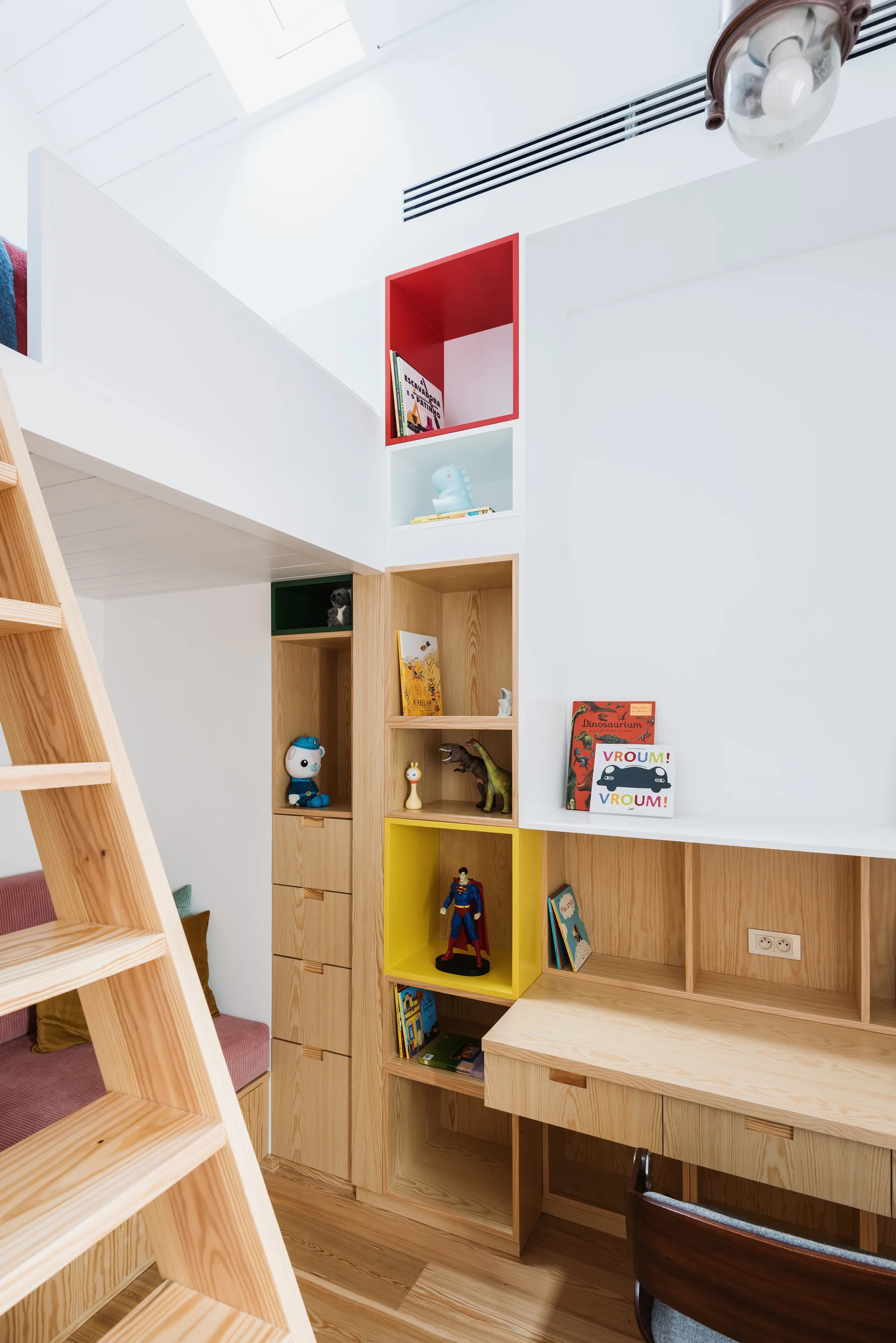 A kid's bedroom featuring a natural wood loft bed, a built-in desk, and built-in shelving with bright red and yellow painted cubbies.