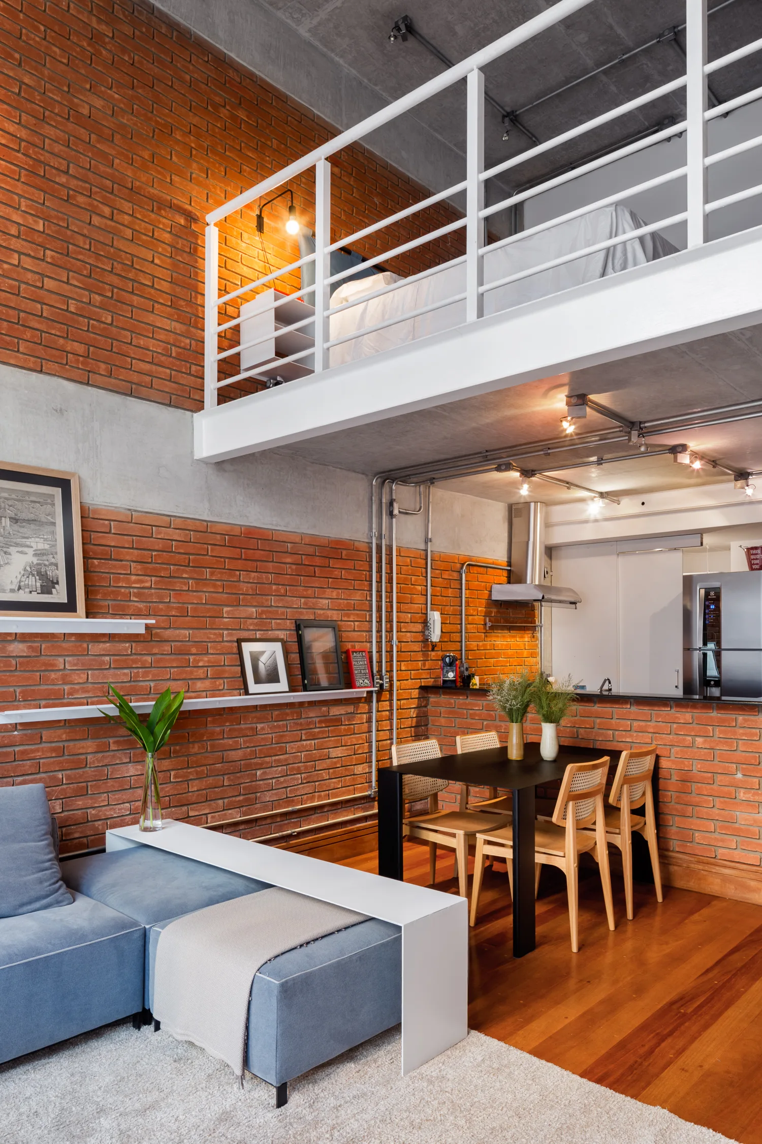 An industrial loft apartment featuring a blue sectional sofa, a black dining table, exposed red brick walls, and a mezzanine level with a bed.