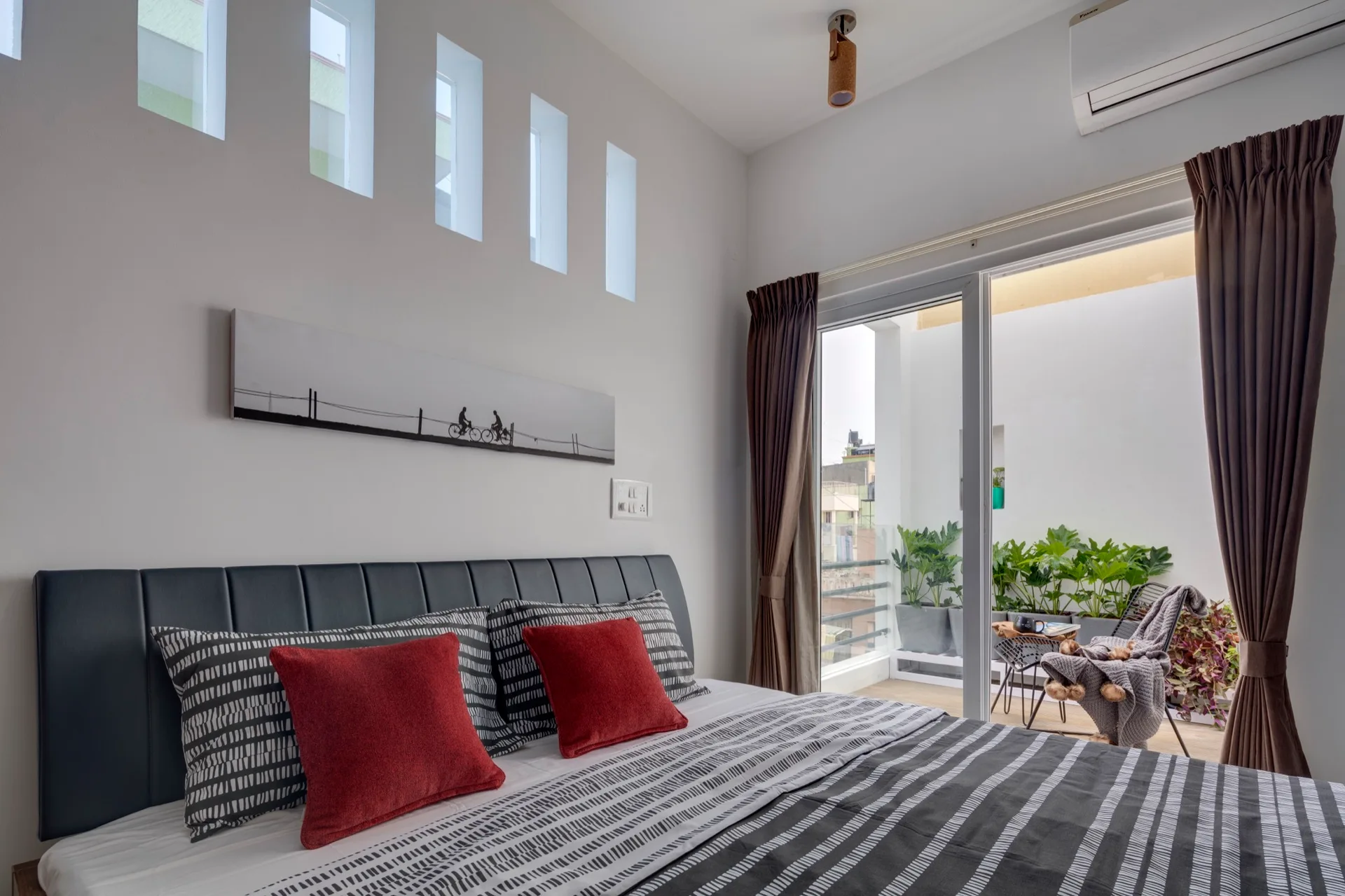 A modern bedroom features a grey upholstered headboard, striped bedding with red pillows, and sliding glass doors opening to a plant-filled balcony.