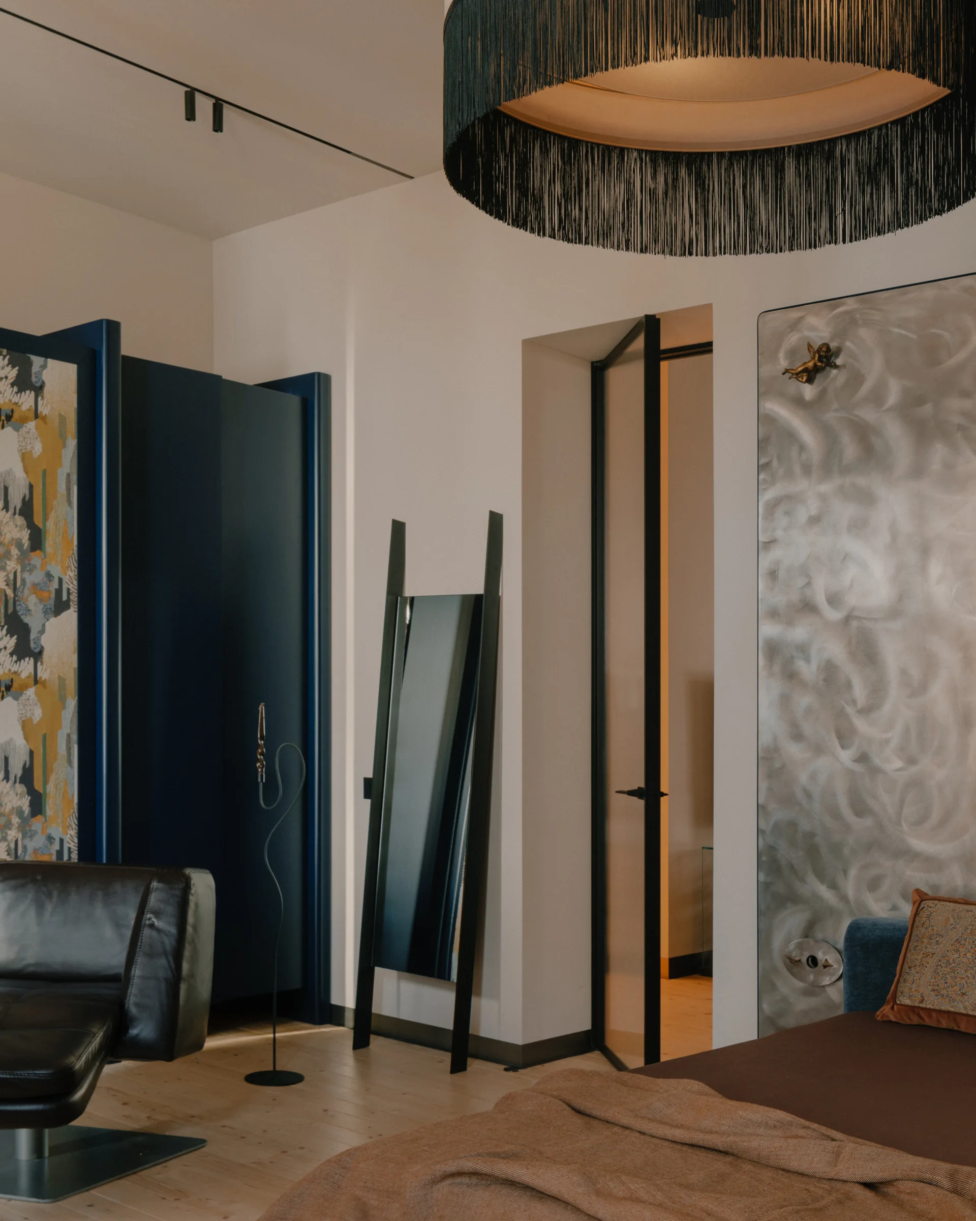 An eclectic bedroom features a large black fringe chandelier hanging above wooden floors near a tall glass door and a blue cabinet.