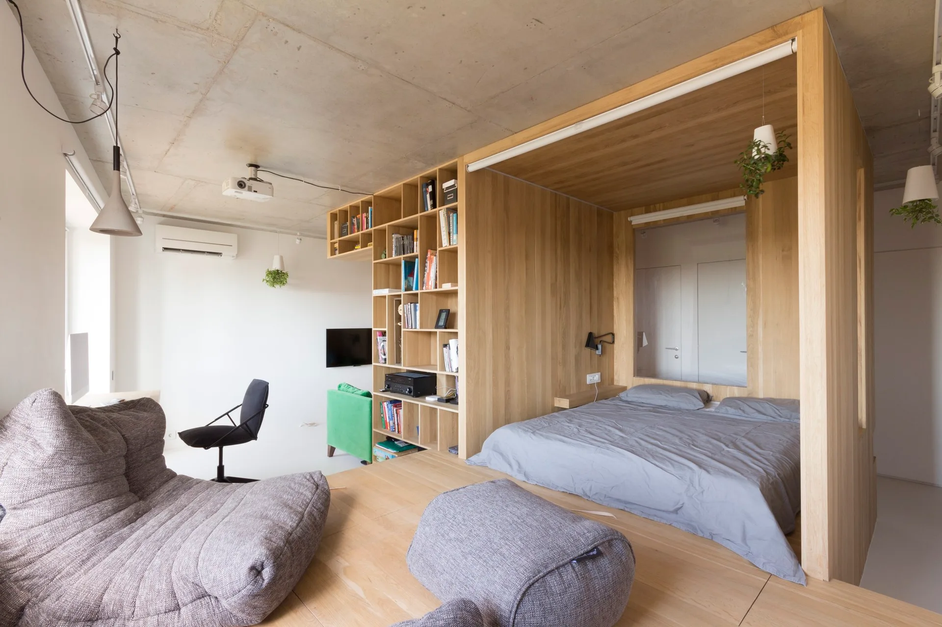 A modern studio apartment features a large light wood structure containing a bed and bookshelf, adjacent to a grey lounge area under a concrete ceiling.