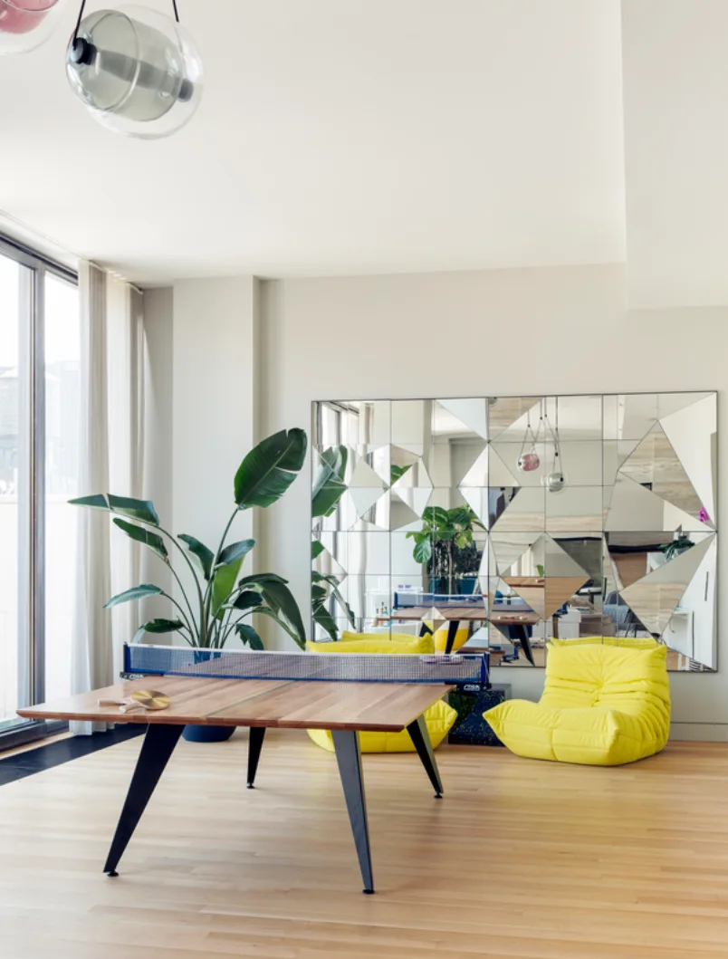 A bright modern recreation room featuring a wooden ping pong table, low-profile yellow lounge chairs, and a large faceted geometric mirror.