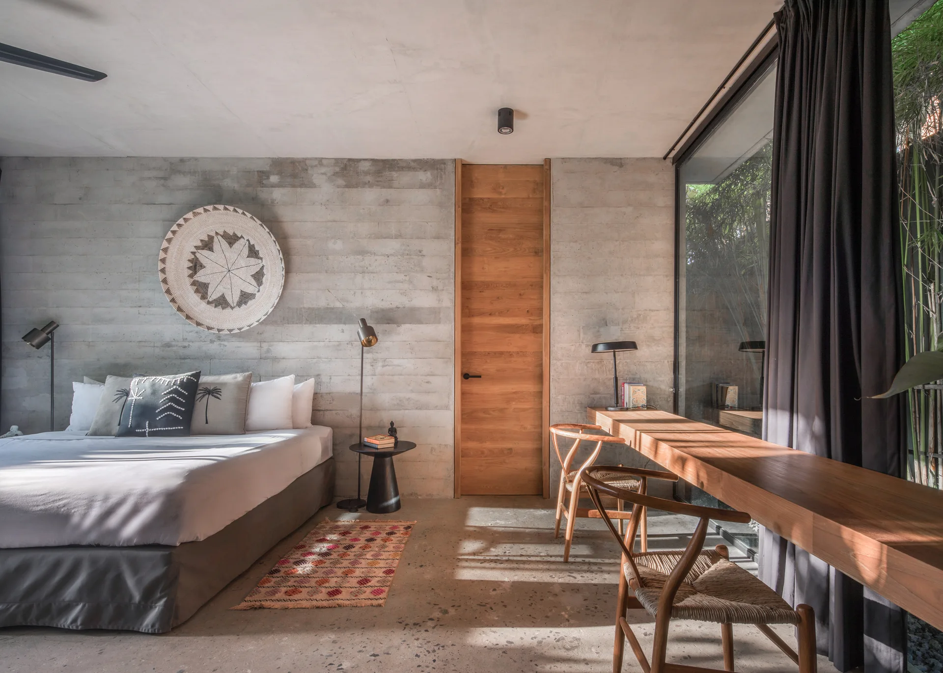A modern bedroom features a bed with grey bedding, raw concrete walls, a full-height wood door, and a built-in wooden desk along a large glass window.