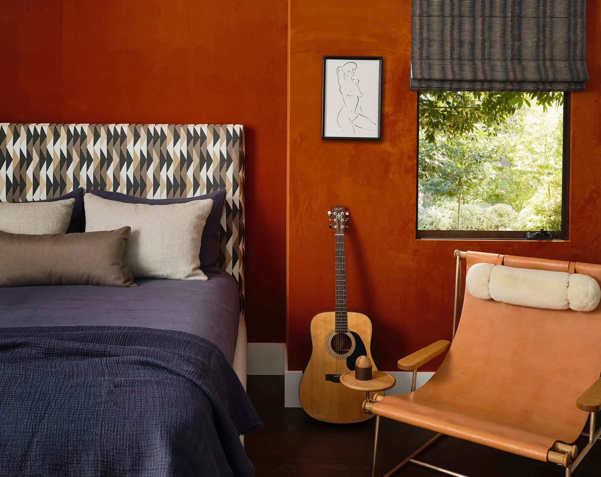 A bedroom corner features a vibrant burnt orange wall, an upholstered geometric headboard with dark bedding, and a leather sling chair next to a guitar.