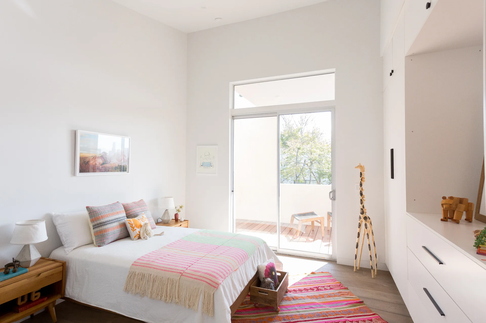 A bright kids' bedroom features a bed with striped bedding, white built-in closets, wood nightstands, and a tall toy giraffe next to a sliding glass door.