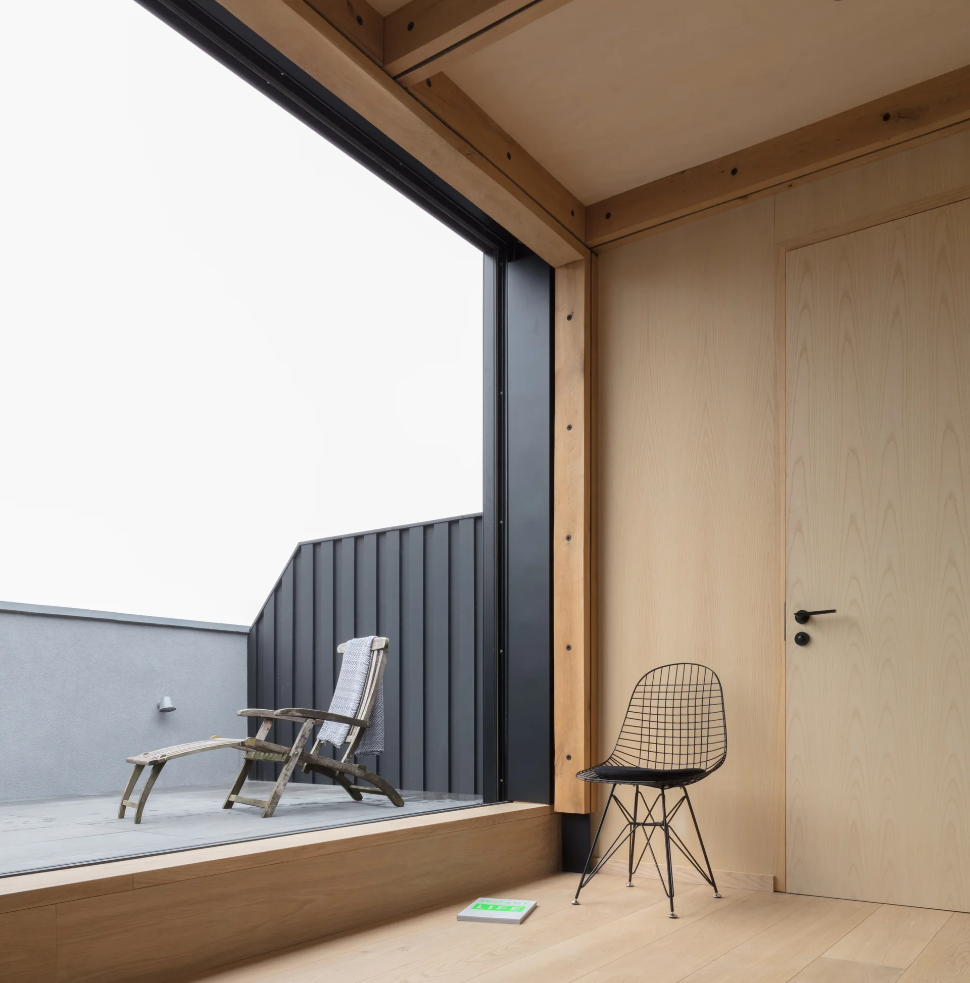 A minimalist light wood paneled room with a black wire chair next to a large sliding glass door opening to a grey terrace with a wooden lounge chair.