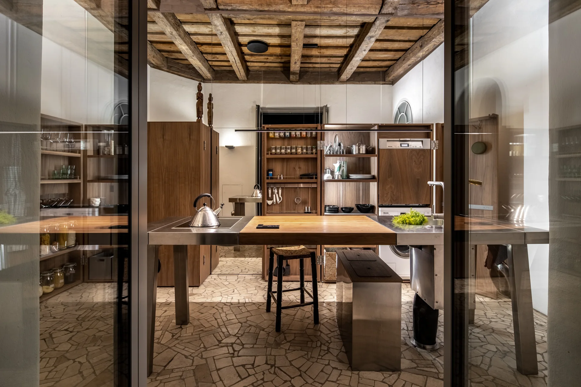 A rustic modern kitchen viewed through glass doors, featuring a stainless steel and wood island, timber ceiling beams, and a flagstone floor.