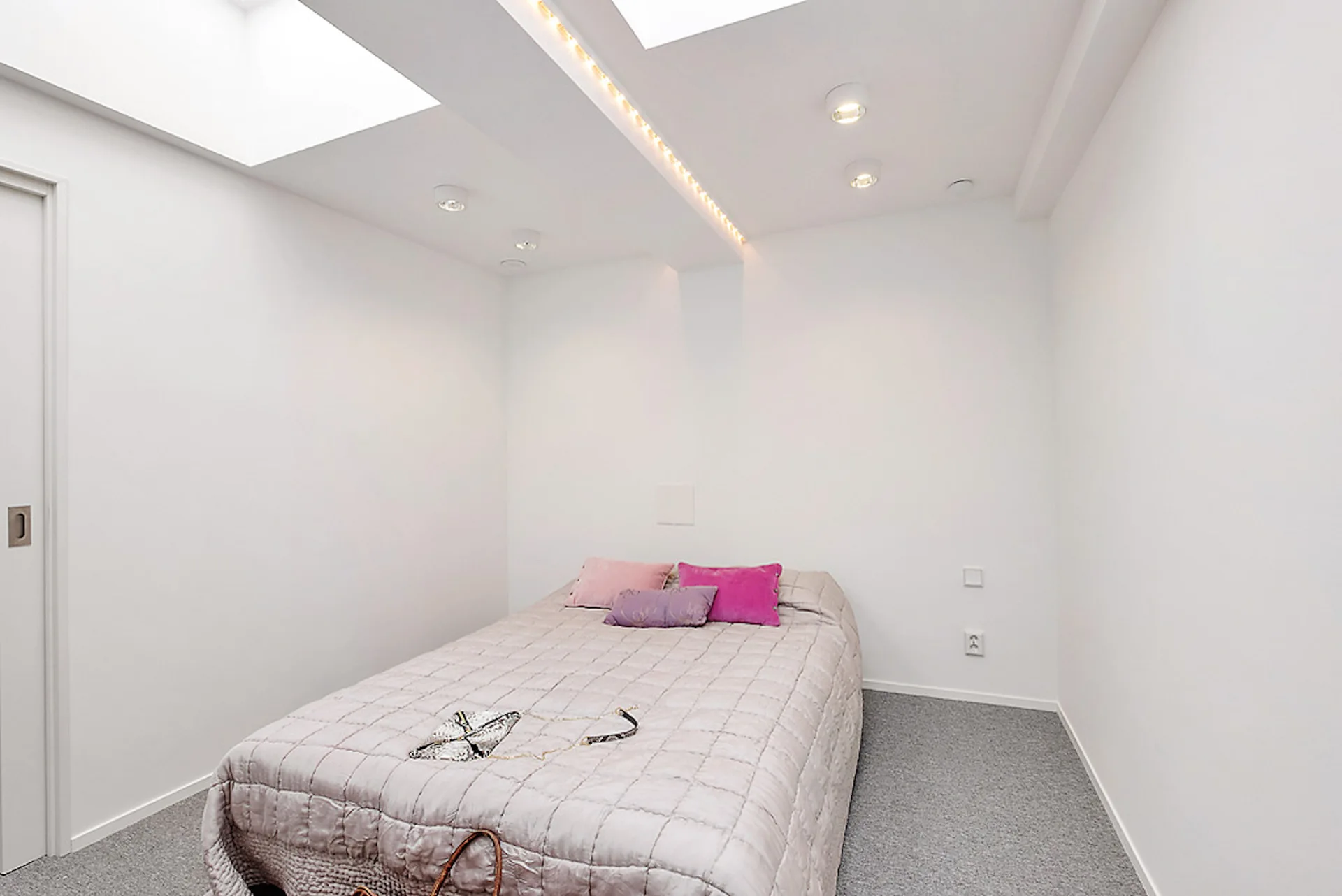 A minimalist white bedroom featuring a low bed with pink throw pillows underneath two large rectangular ceiling skylights.