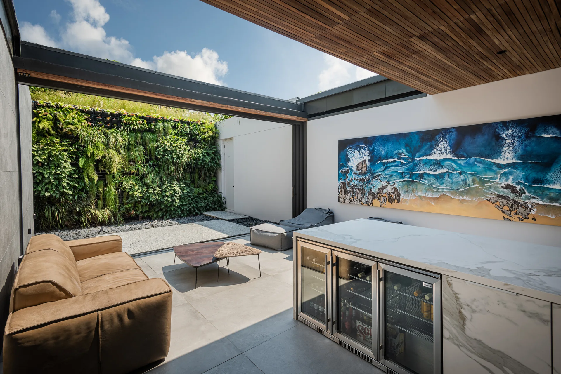 A modern outdoor patio features a tan leather sofa, a marble bar with beverage refrigerators, and a lush vertical garden wall.