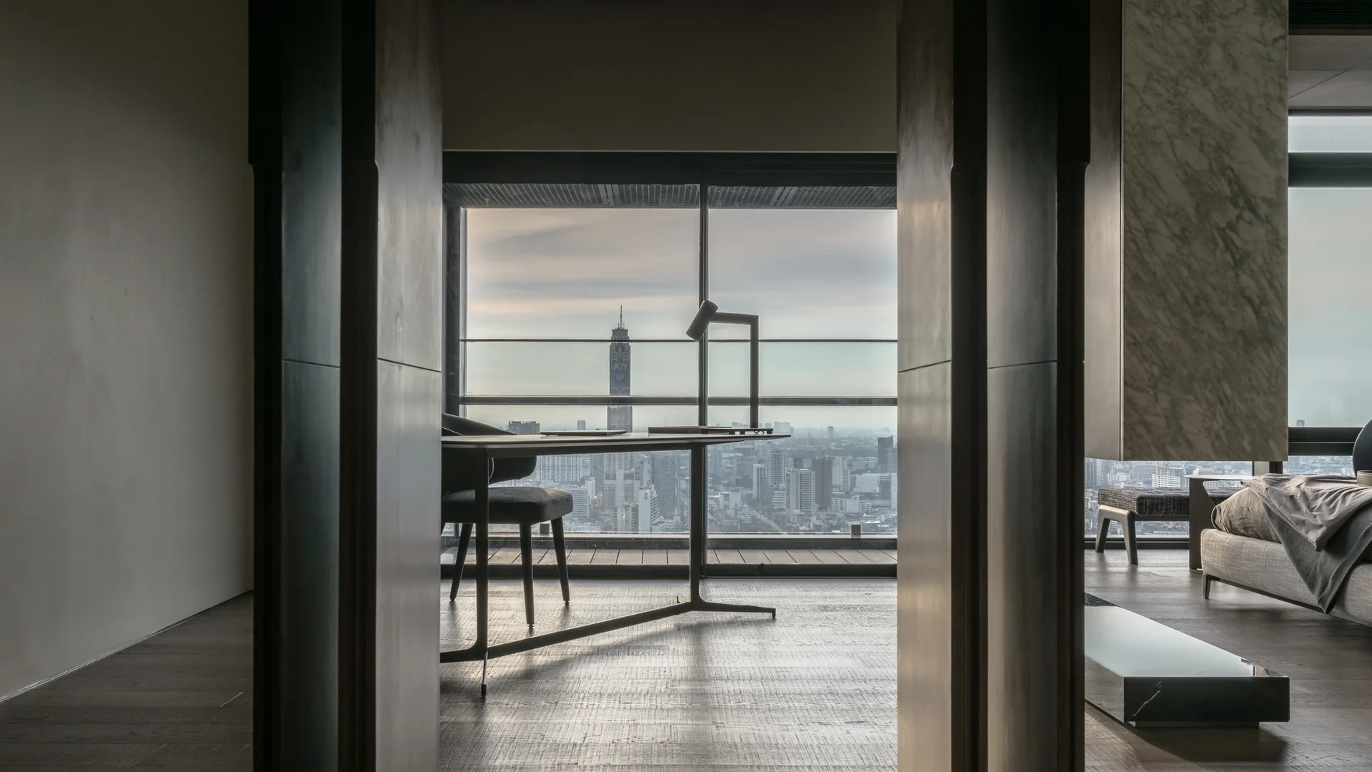 A minimalist home office framed by dark wood paneling features a slim desk, small desk lamp, and floor-to-ceiling window overlooking a city skyline.