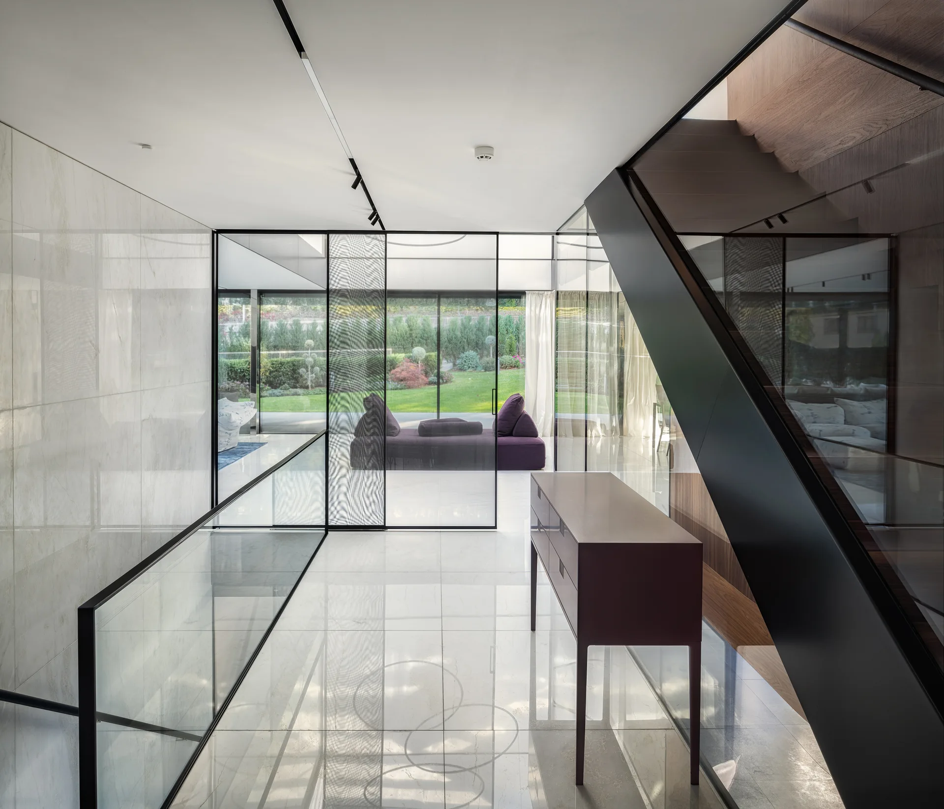 A modern foyer featuring polished marble floors, an angular black staircase, a slim console table, and a glass partition looking into a garden room.