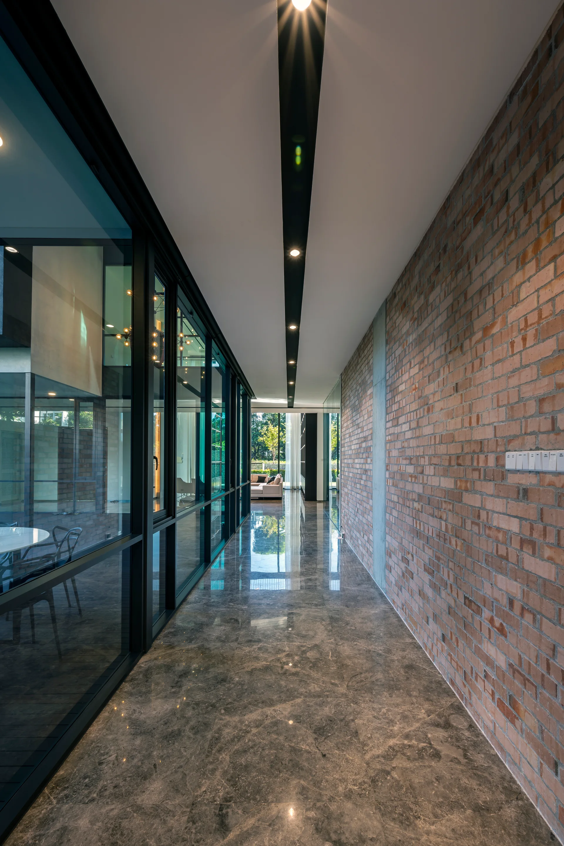 A long modern hallway featuring an exposed red brick wall, a floor-to-ceiling glass partition, polished grey stone flooring, and recessed ceiling lights.