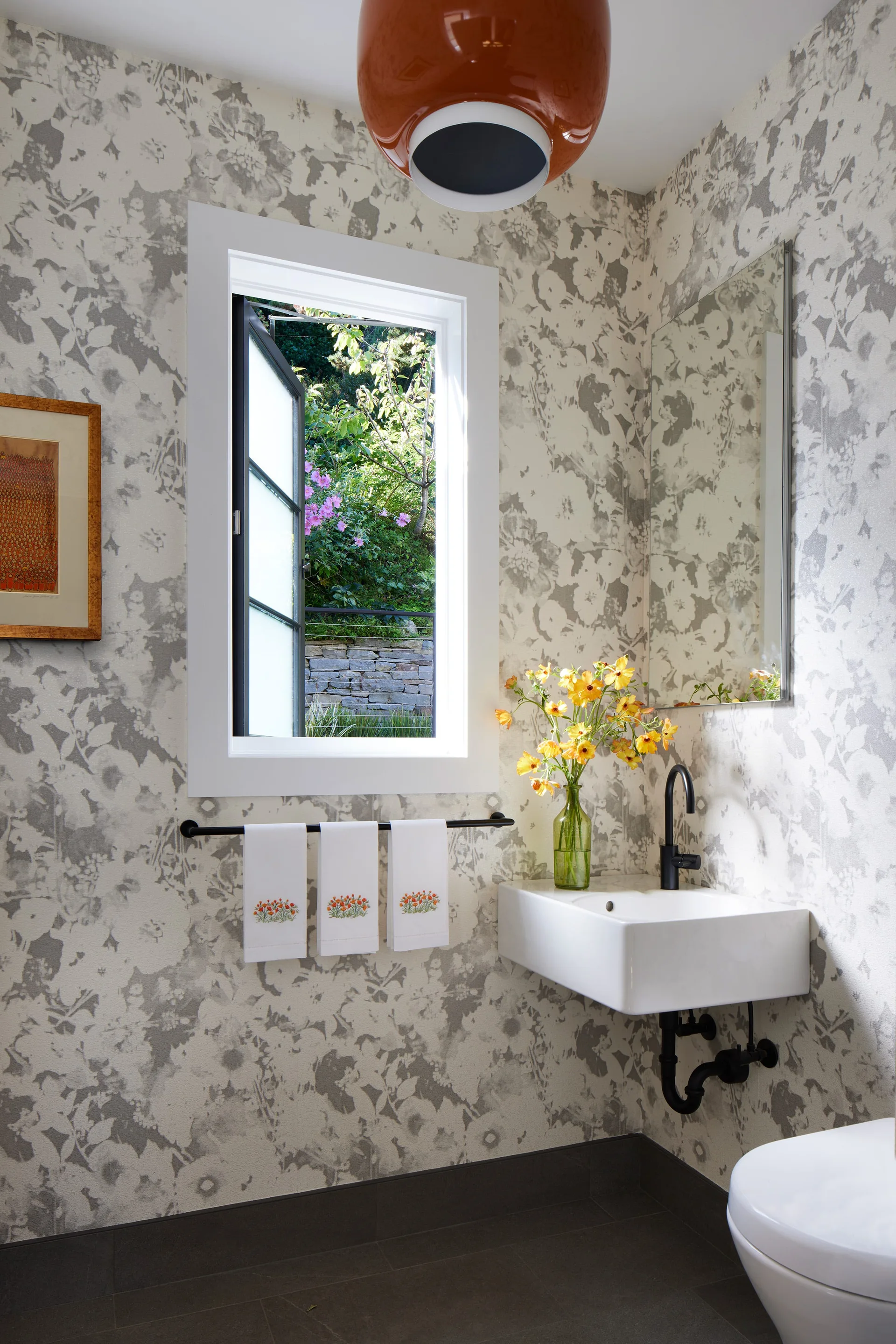 A modern powder room with grey floral wallpaper, a white floating sink with black fixtures, and a glossy orange pendant light.