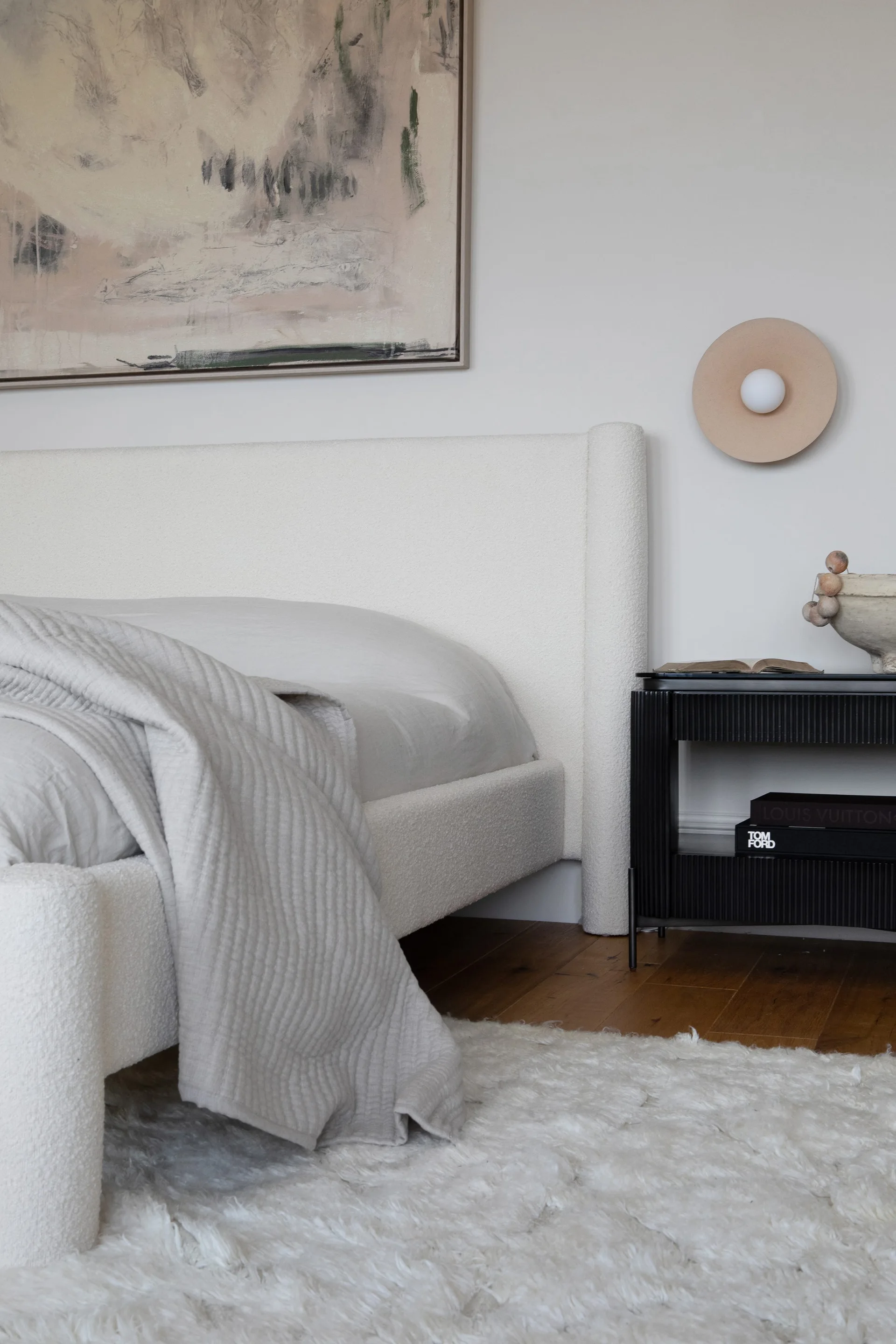 A neutral bedroom features a textured cream bouclé bed, a black fluted wooden nightstand, a white shag rug, and an abstract painting.
