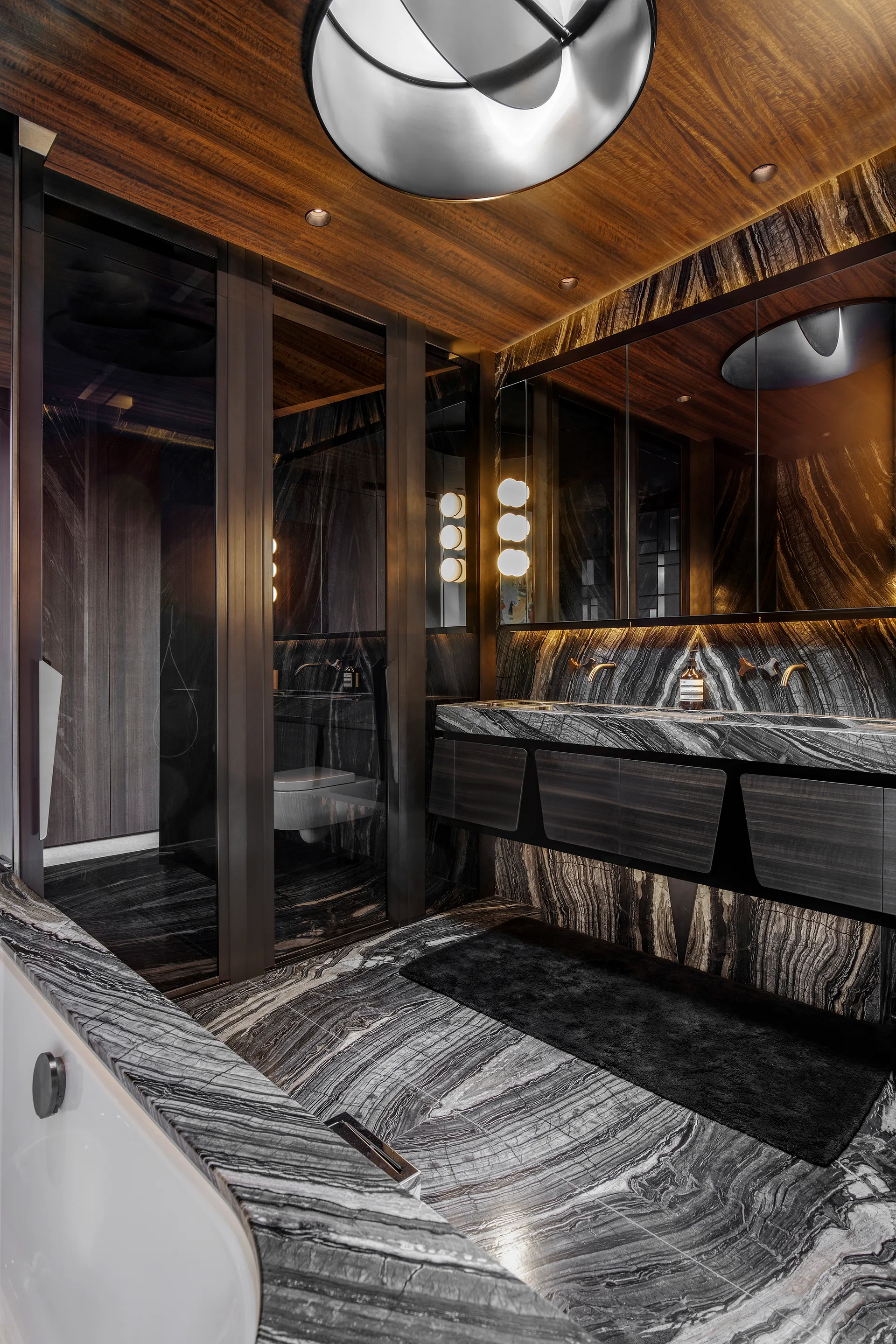 A modern bathroom features dark veined marble walls and floors, a warm wood plank ceiling, a floating vanity with gold faucets, and a large mirror.
