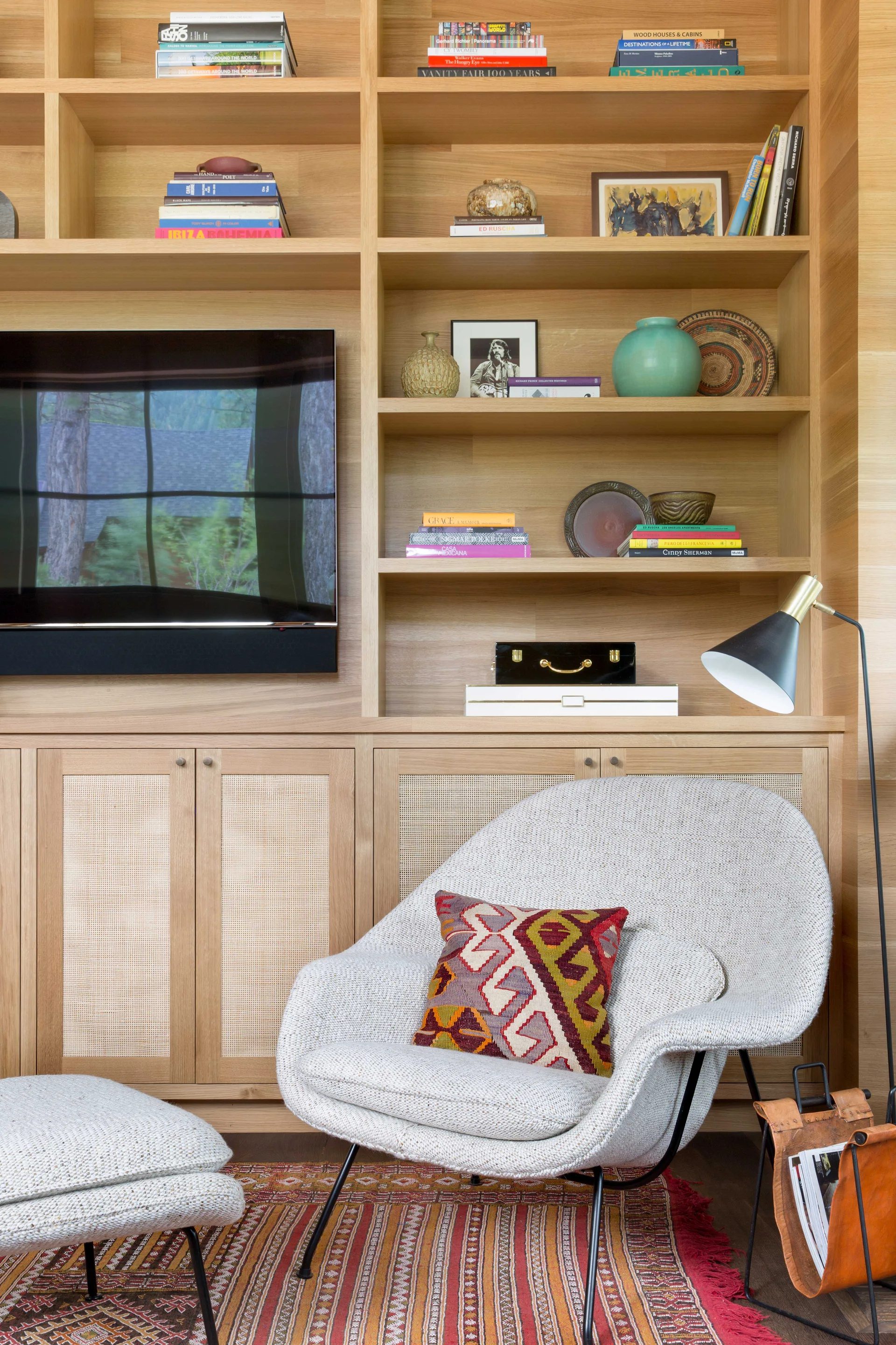 A textured white lounge chair with a patterned pillow sits before a wood built-in bookcase featuring cane panel cabinets and a television.